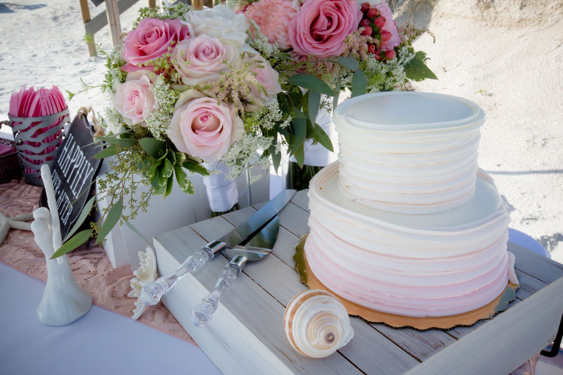 Wedding cake and floral arrangement on beach, pink and white hues.