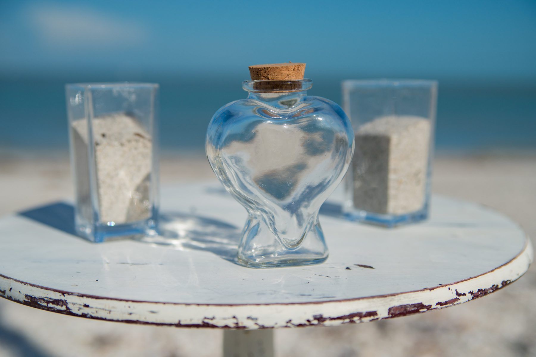 Sand ceremony setup on a white table at the beach: heart-shaped bottle, two glass containers filled with sand, ocean background.