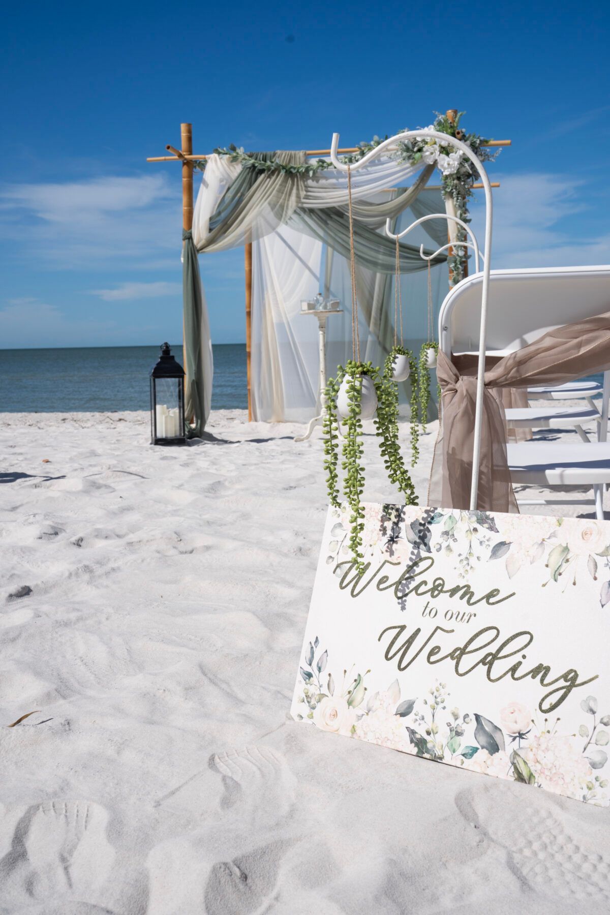 Beach wedding ceremony setup with white chairs, arch decorated with greenery and sheer fabric. 