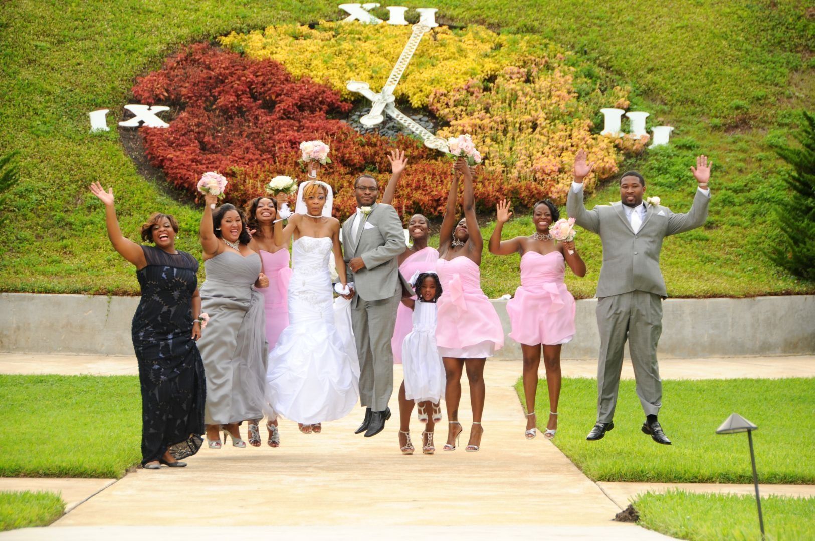 Wedding party jumping in front of floral clock display. People are dressed in formal attire; smiles.