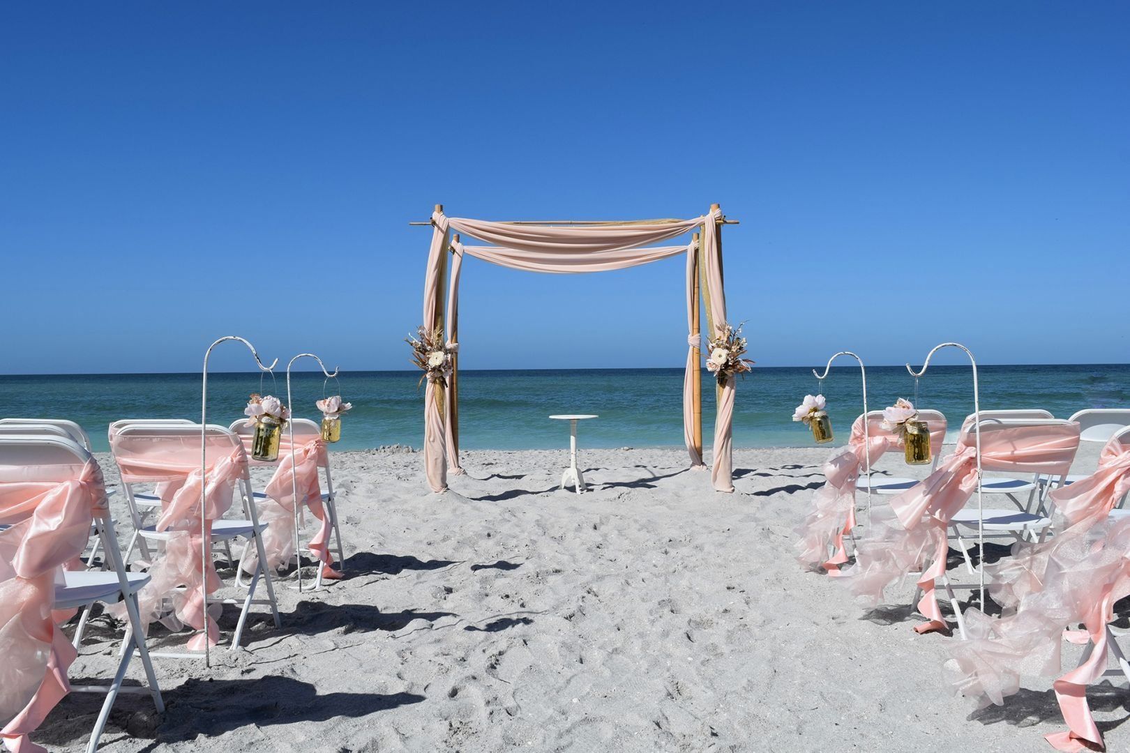 Beach wedding setup with draped arch, chairs with pink ribbons, and ocean view.