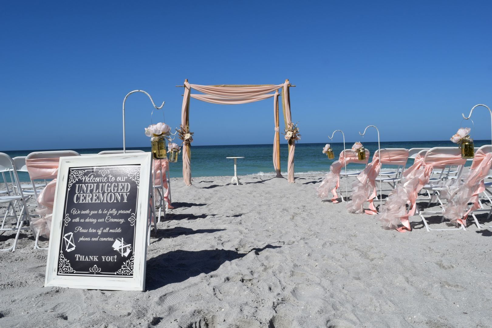 Beach wedding ceremony setup with white chairs, archway, and sign on a sunny day.