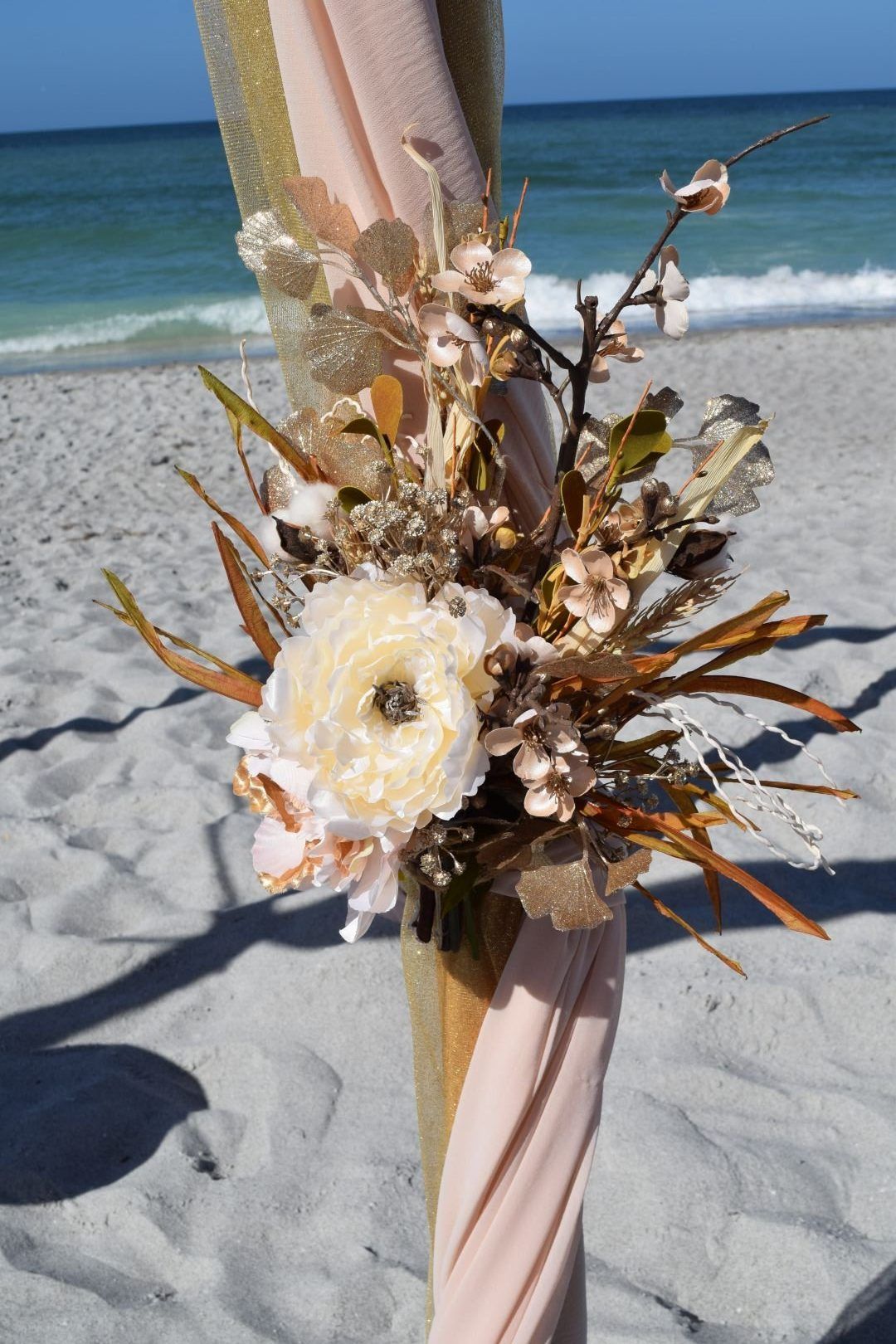 Floral arrangement on draped wedding arch on a sandy beach; ocean in the background.