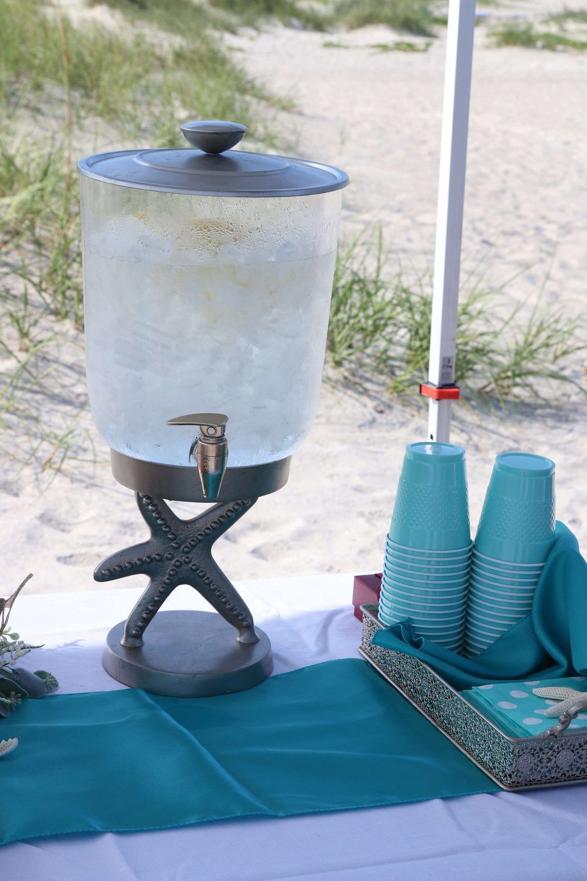 Beverage dispenser on beach table; turquoise cups and napkins, white sand backdrop.