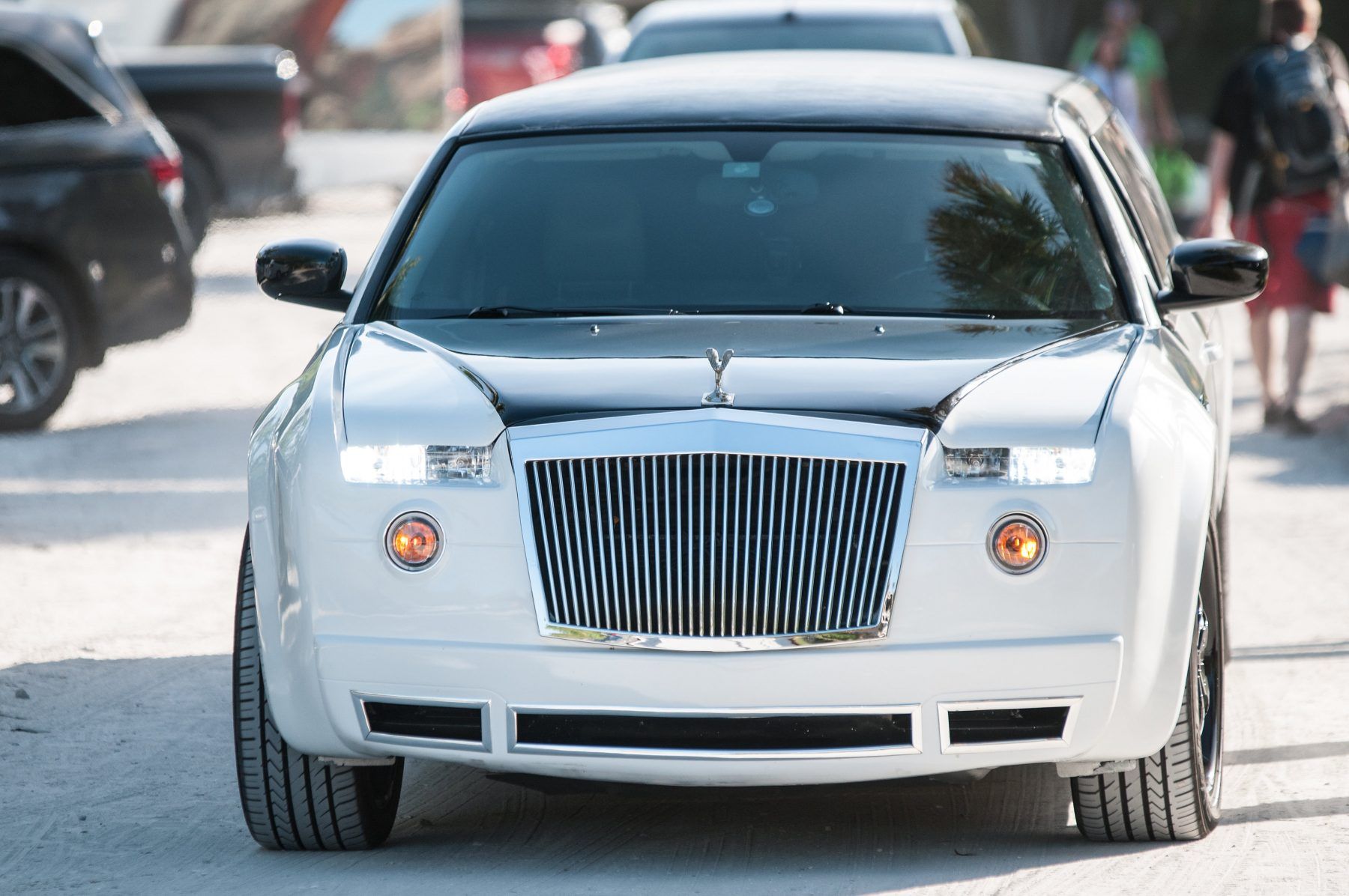 White and black car with a large grille, resembling a Rolls Royce, on a road.