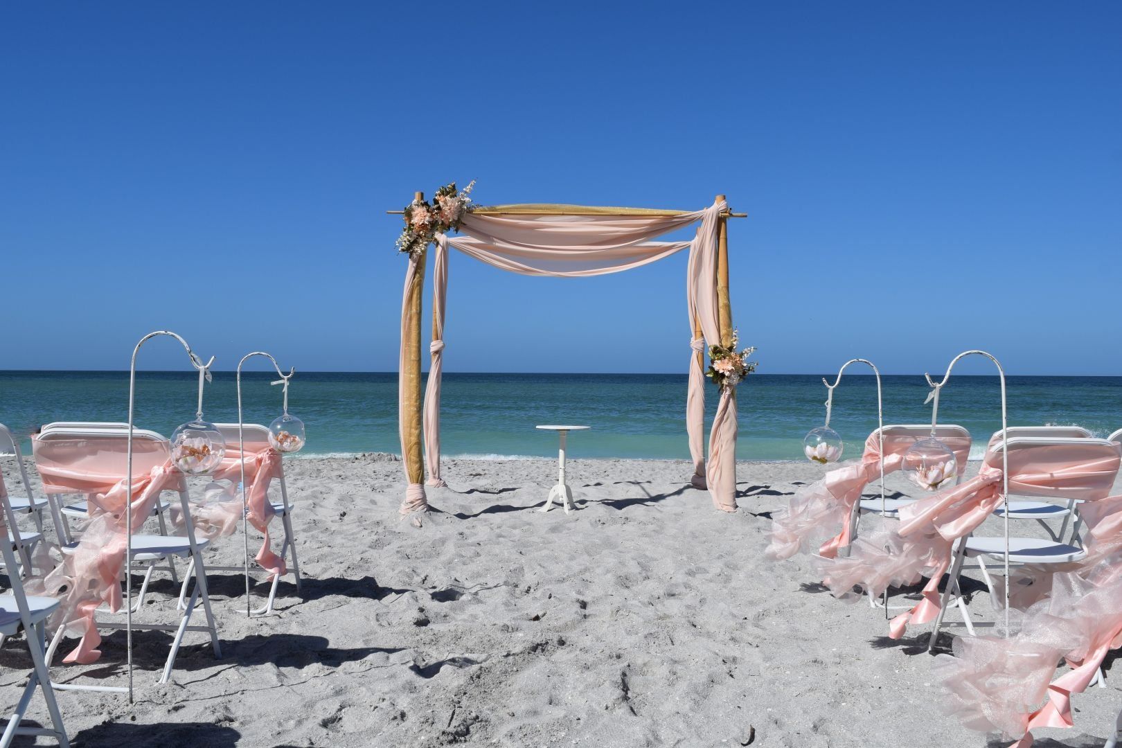 Beach wedding setup: draped arch, white chairs with pink accents, ocean and blue sky background.