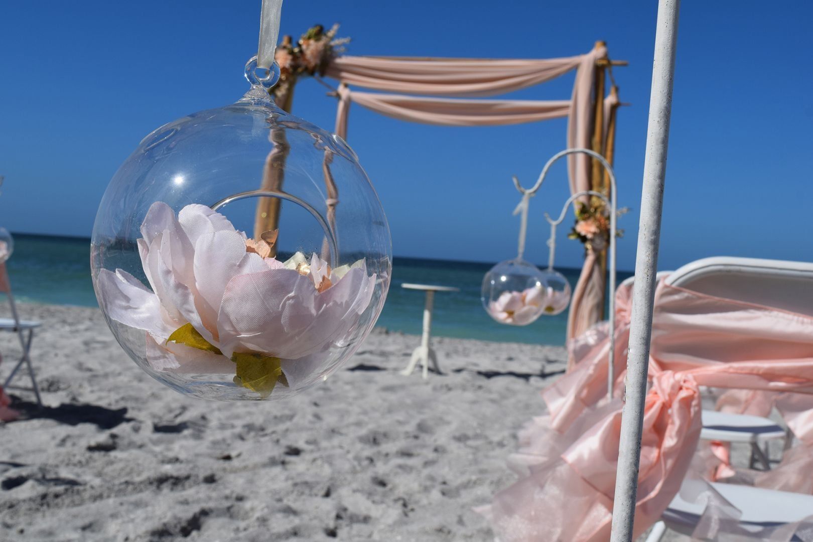 Wedding decor on a beach. Clear globes with flowers hang from a white structure near chairs and a draped arch.