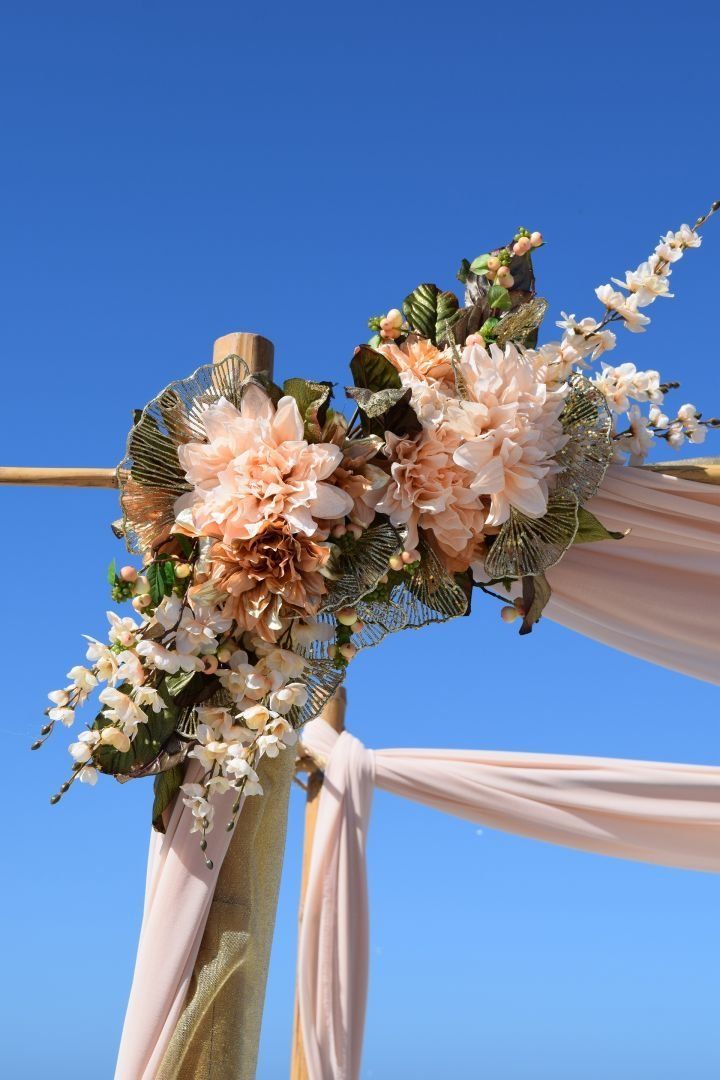 Floral arrangement with peach dahlias and white flowers on a wedding arch with draped fabric against a blue sky.