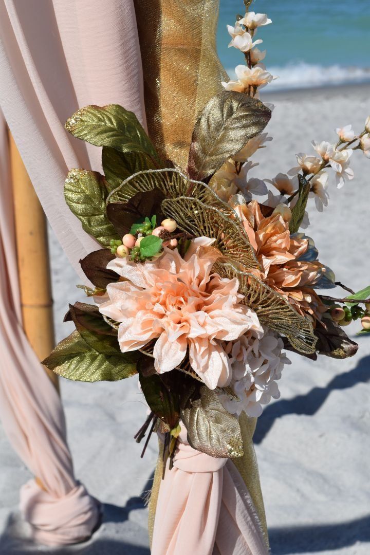 Floral arrangement on bamboo pole with peach and gold accents at beach wedding.
