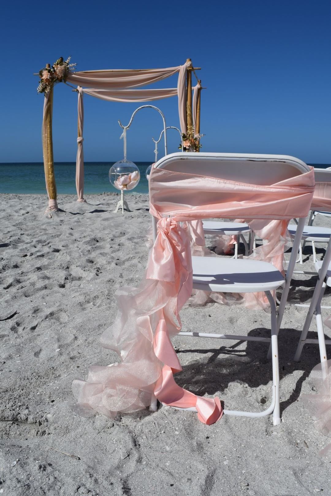 Beach wedding setup with chairs draped in pink fabric, archway, and clear blue sky.