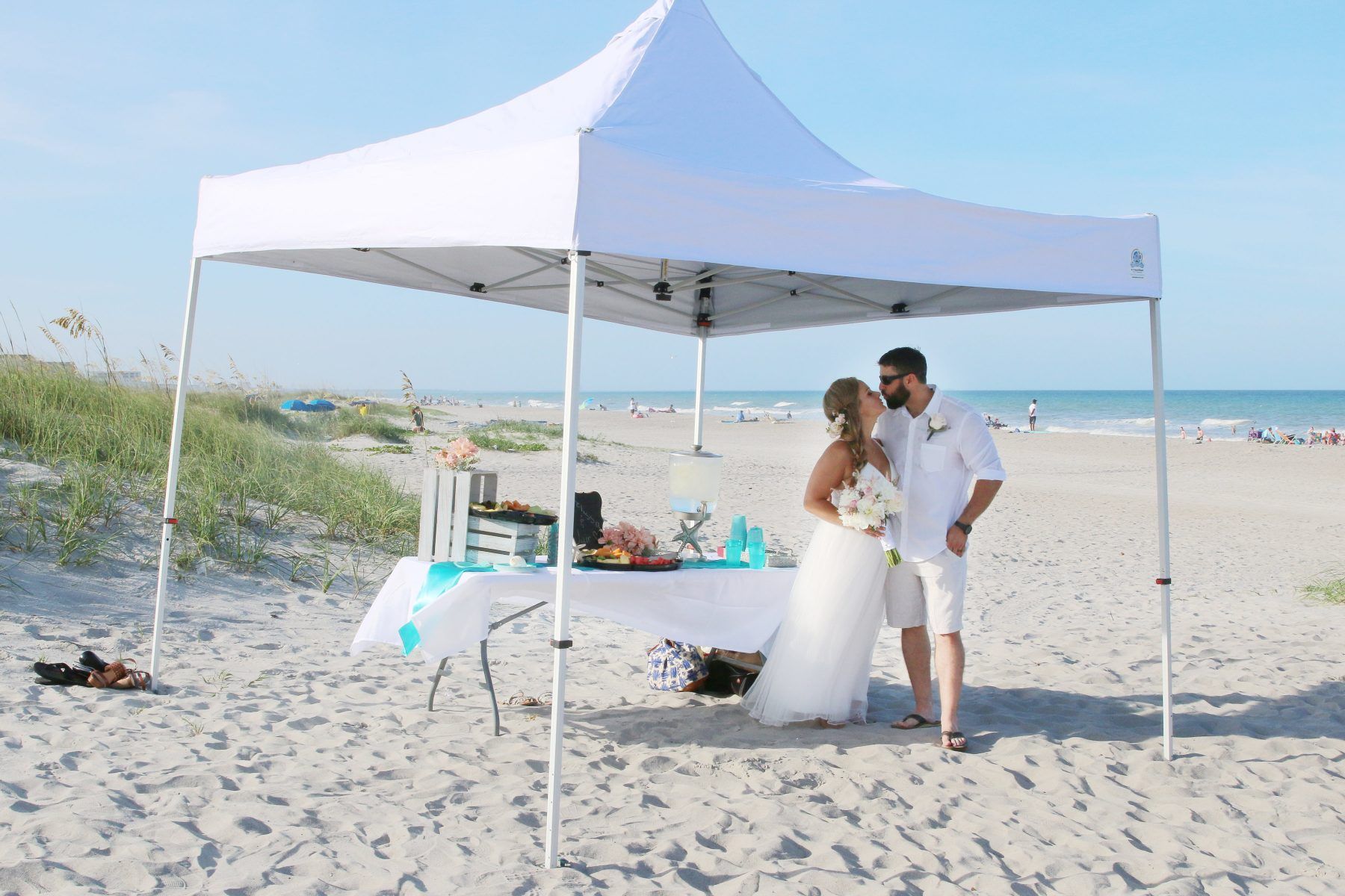 Bride and groom under a white tent on a beach, embracing. A table with decorations is present.