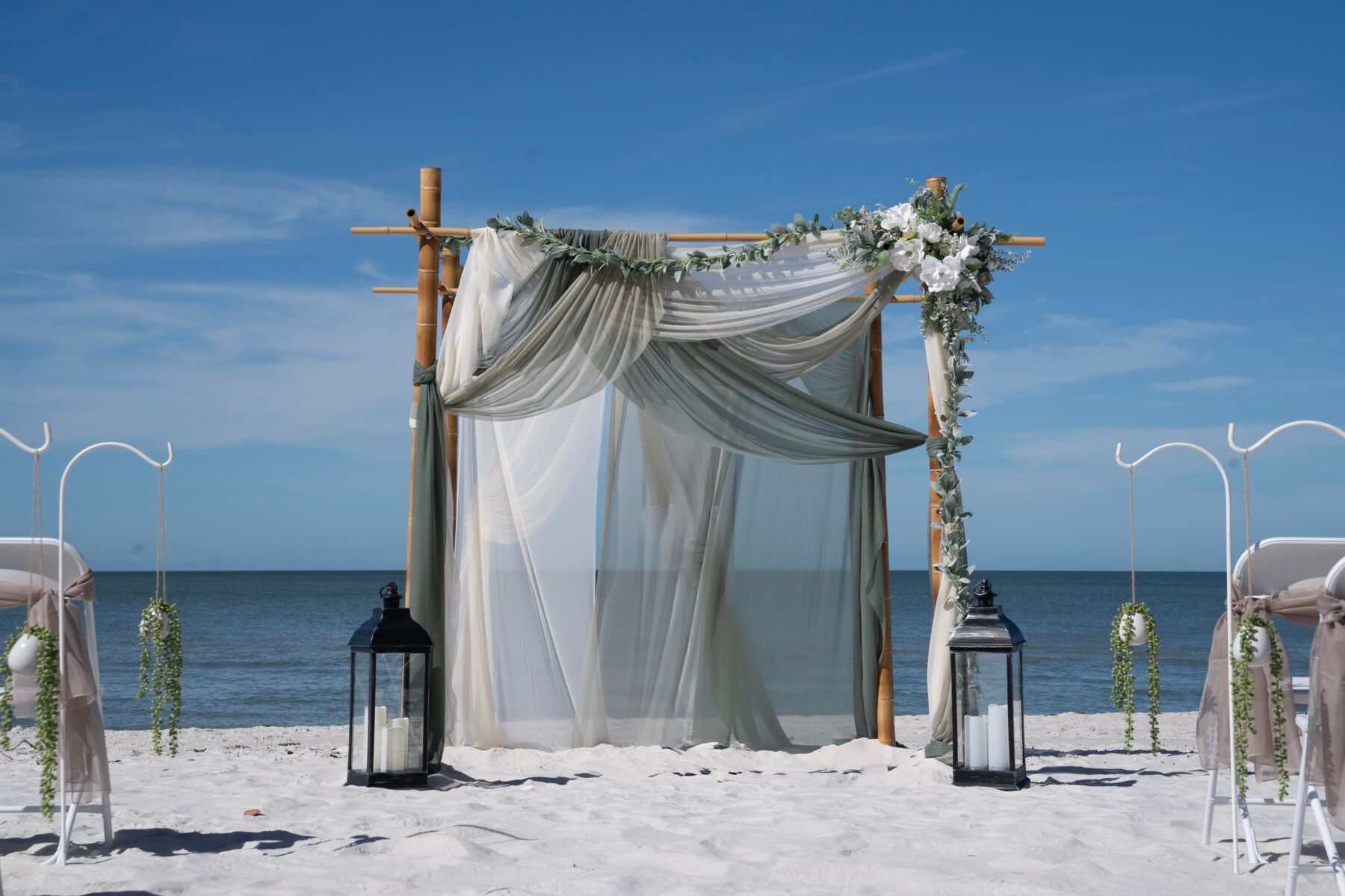 Beach wedding setup with draped fabric canopy, floral accents, lanterns, and ocean backdrop.
