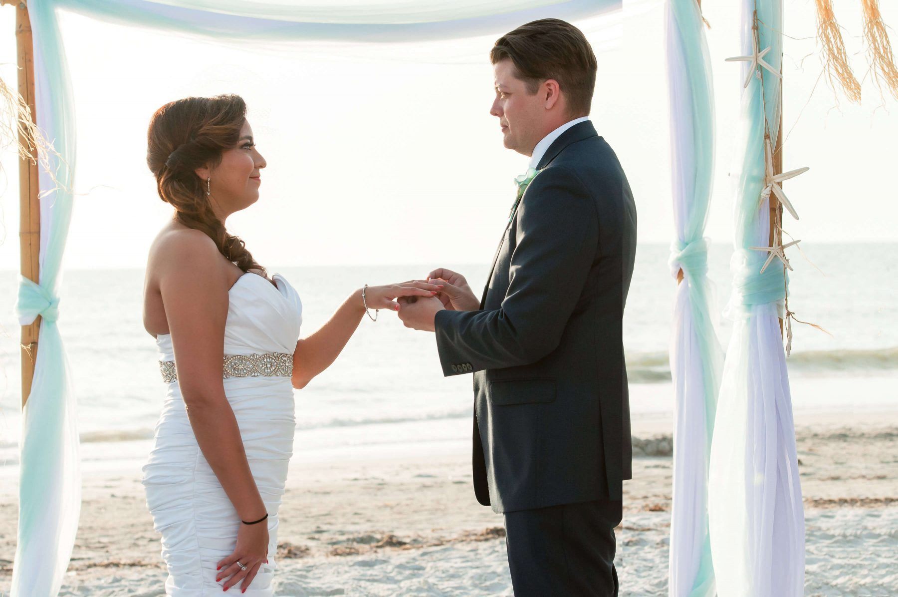 Groom placing ring on bride's finger during a beach wedding ceremony, sunlight, ocean backdrop.