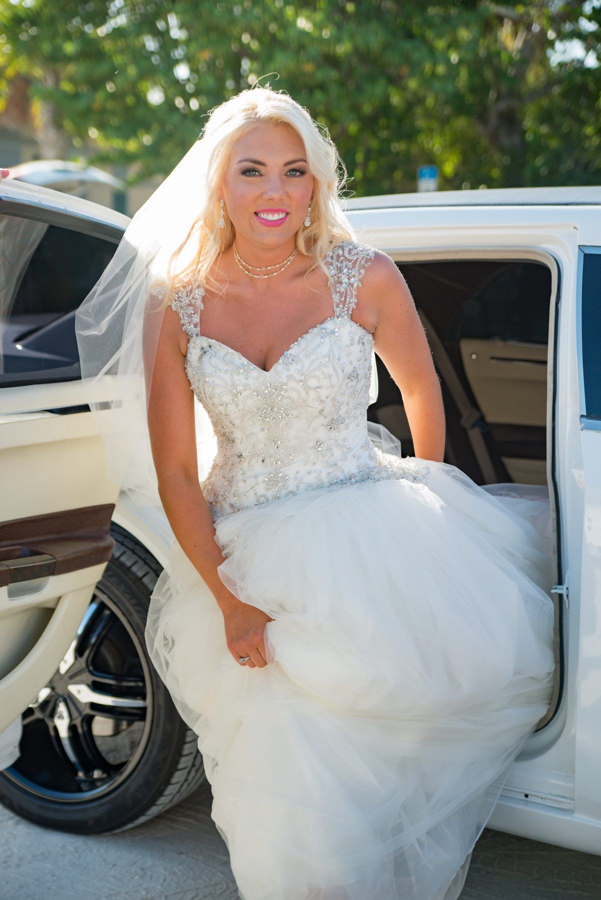 Bride in a white gown exiting a limousine; veil, blonde hair, and smiling.
