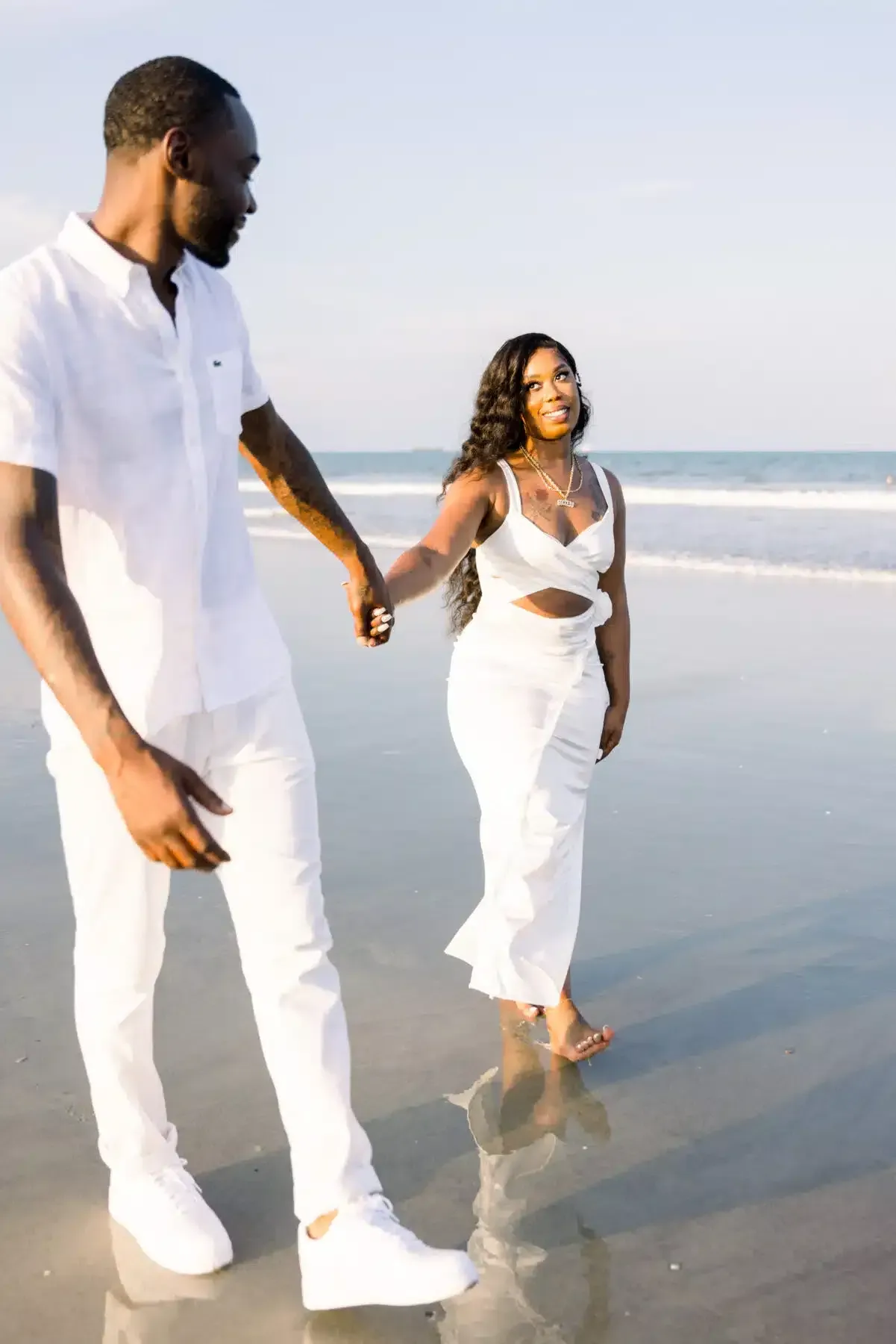 Couple holding hands, walking along a beach in white outfits, smiling, ocean in background.
