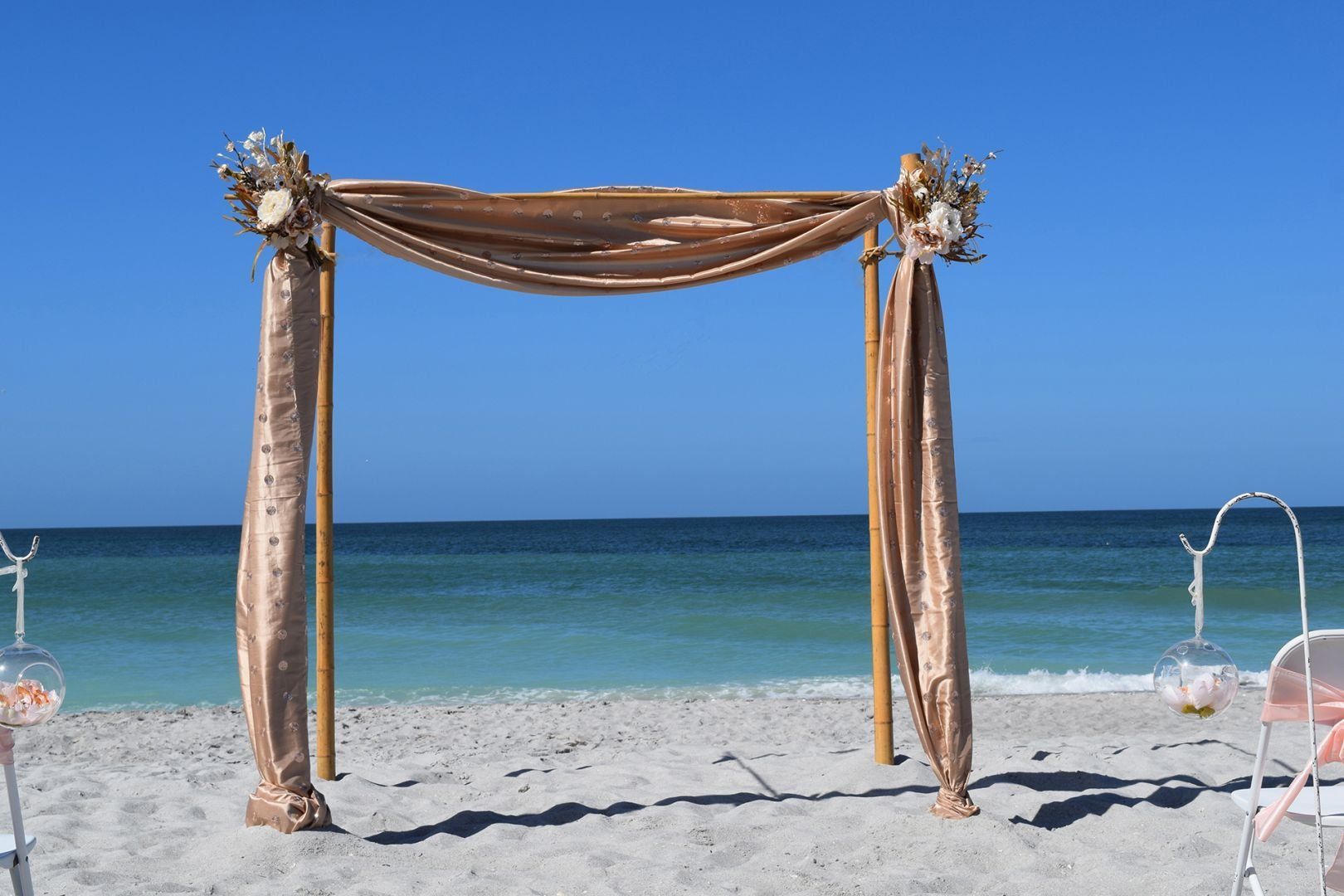 Beach wedding arch with gold fabric, flowers, and ocean view.