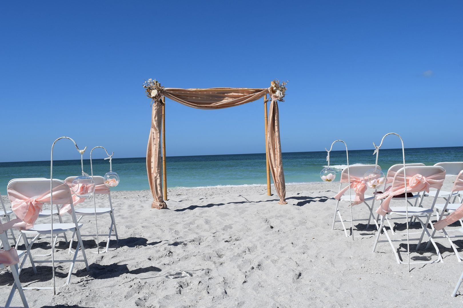 Beach wedding setup: white chairs with pink ribbons face a gold draped arch, ocean backdrop.