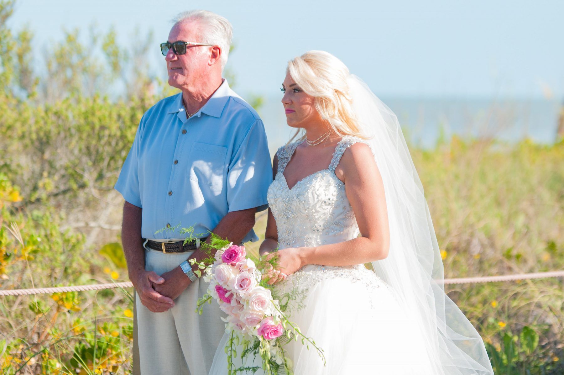 Bride and father at a beach wedding ceremony, father in blue shirt, bride in white dress, holding flowers, looking forward.
