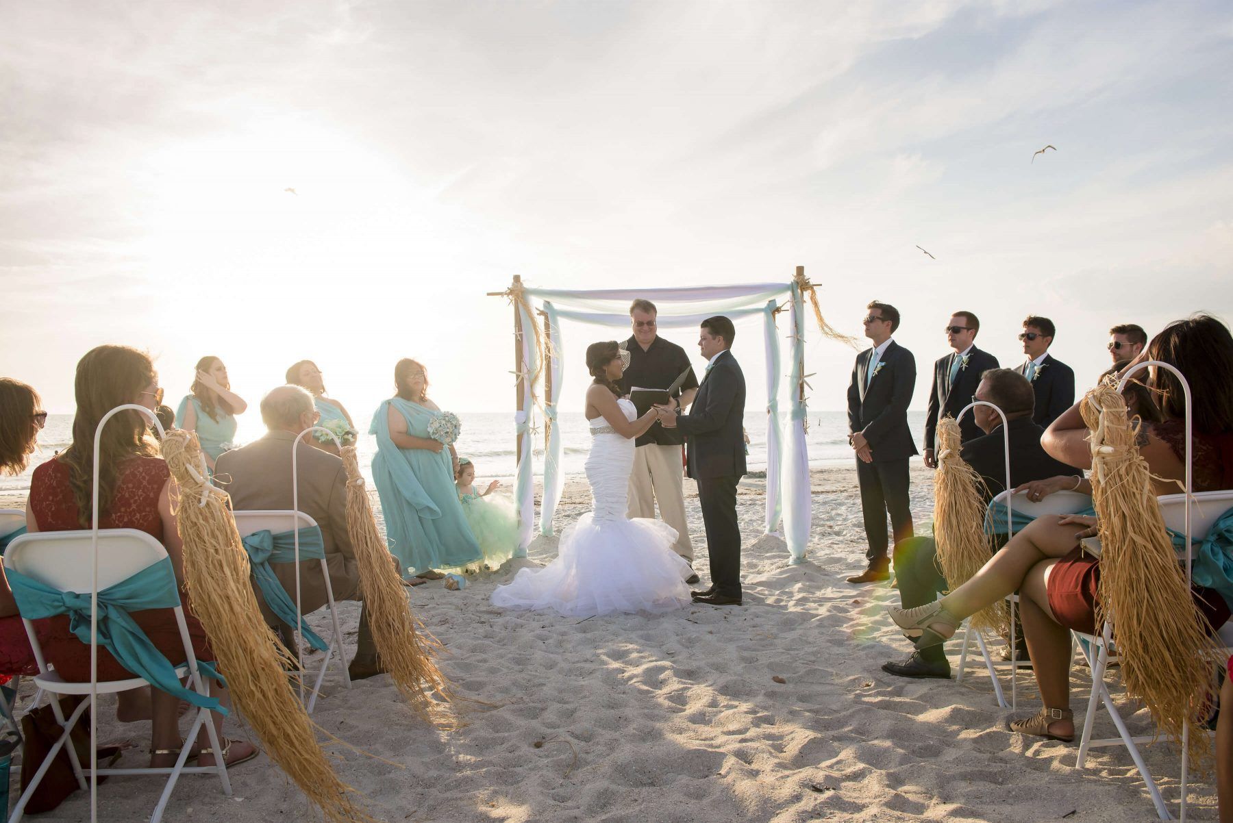 Wedding ceremony on a beach. Bride and groom exchange vows under a decorated arch, guests watch.