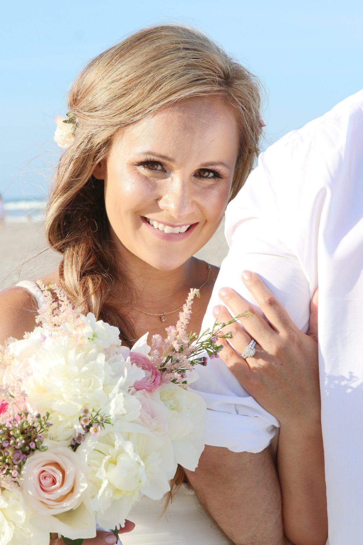 Bride smiling at the camera holding flowers on the beach, with partner's arm visible.