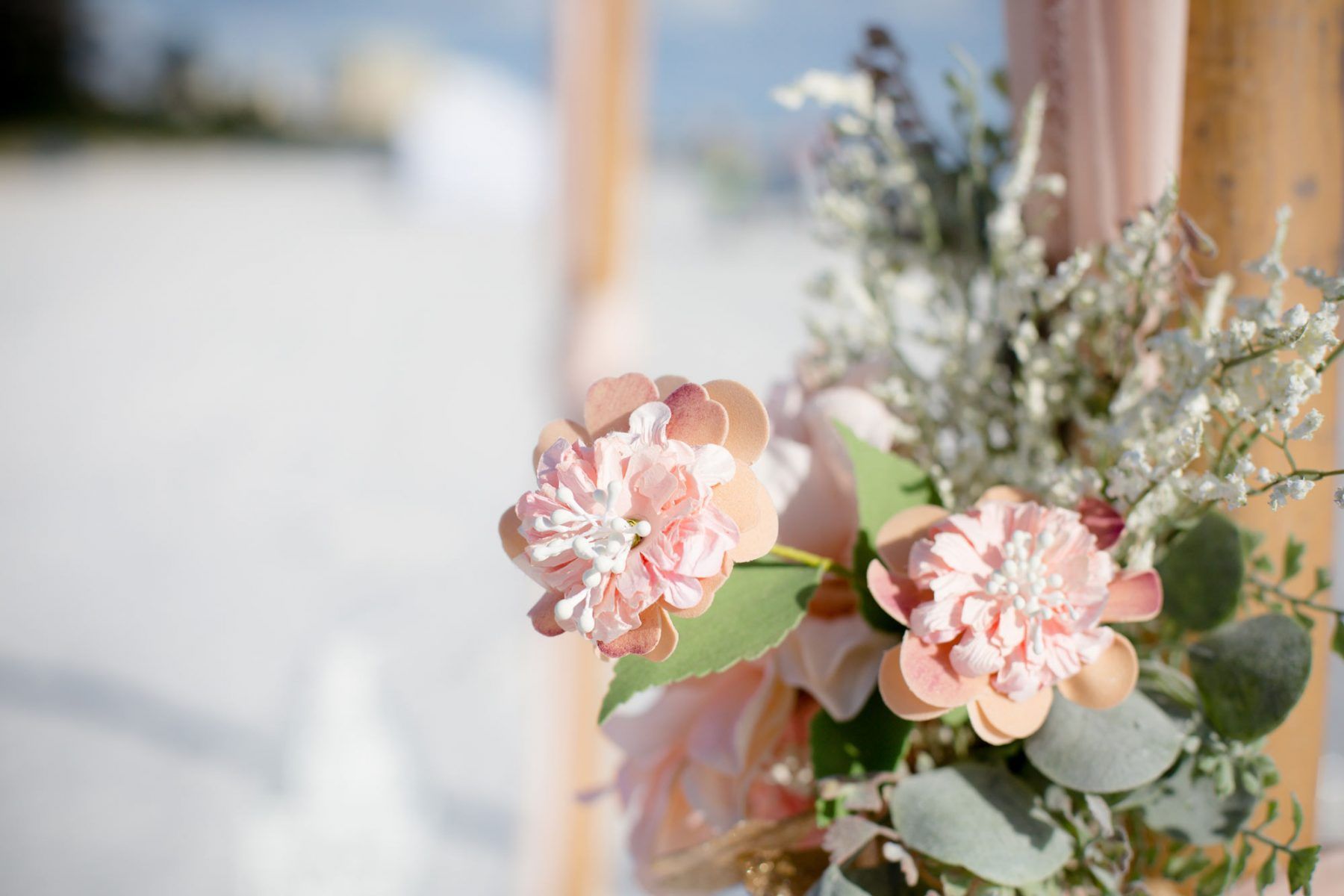 Peach flowers and greenery decorate a wooden wedding arch on a sandy beach.