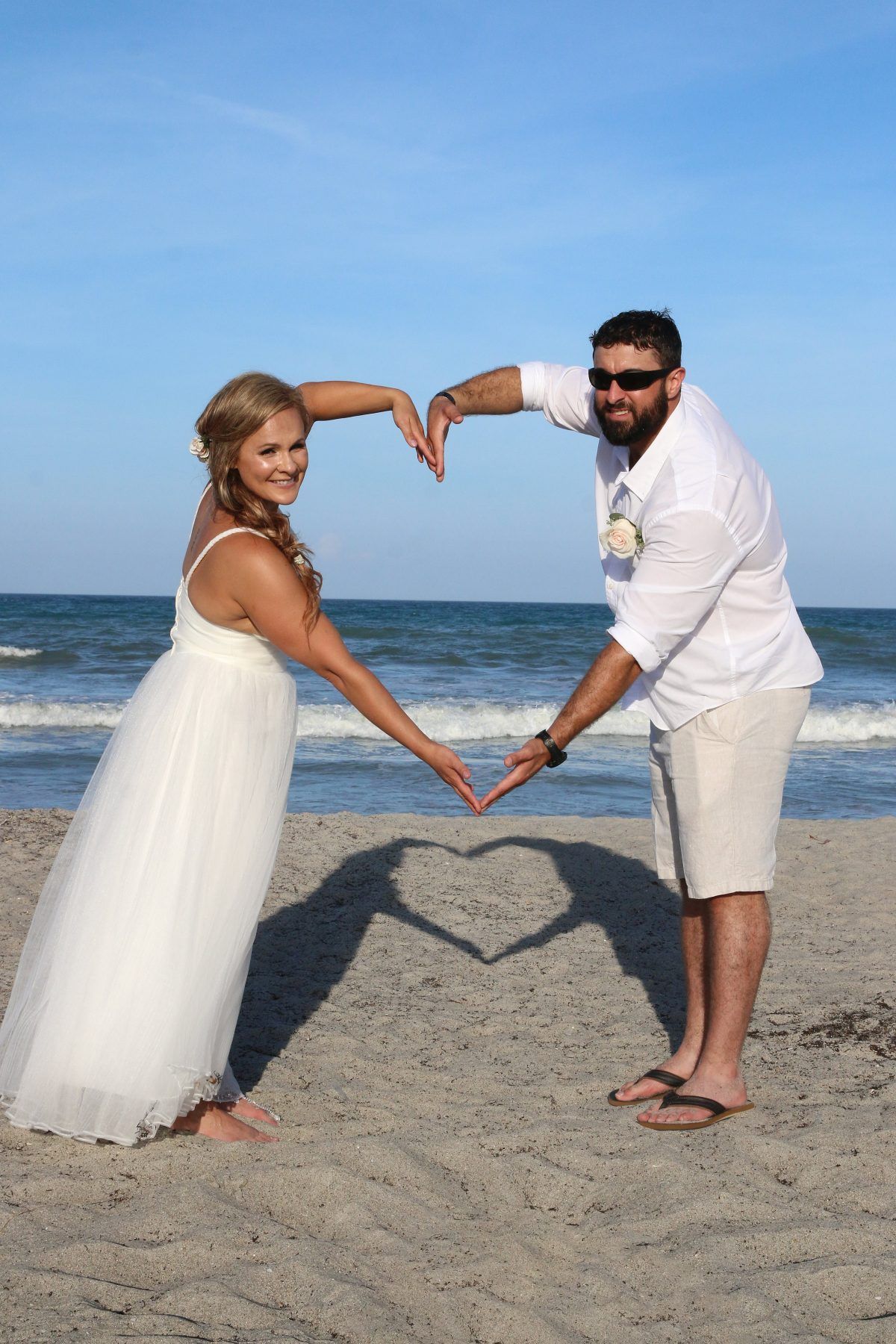 Bride and groom making heart shape with arms on beach, shadow forms heart on sand.