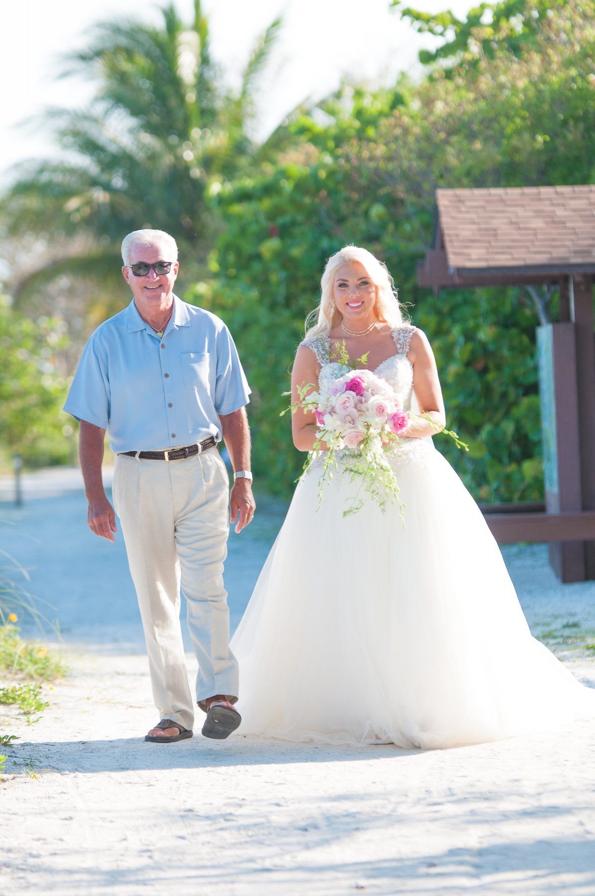 Bride in white gown walks with man in blue shirt and khaki pants on a sandy path.