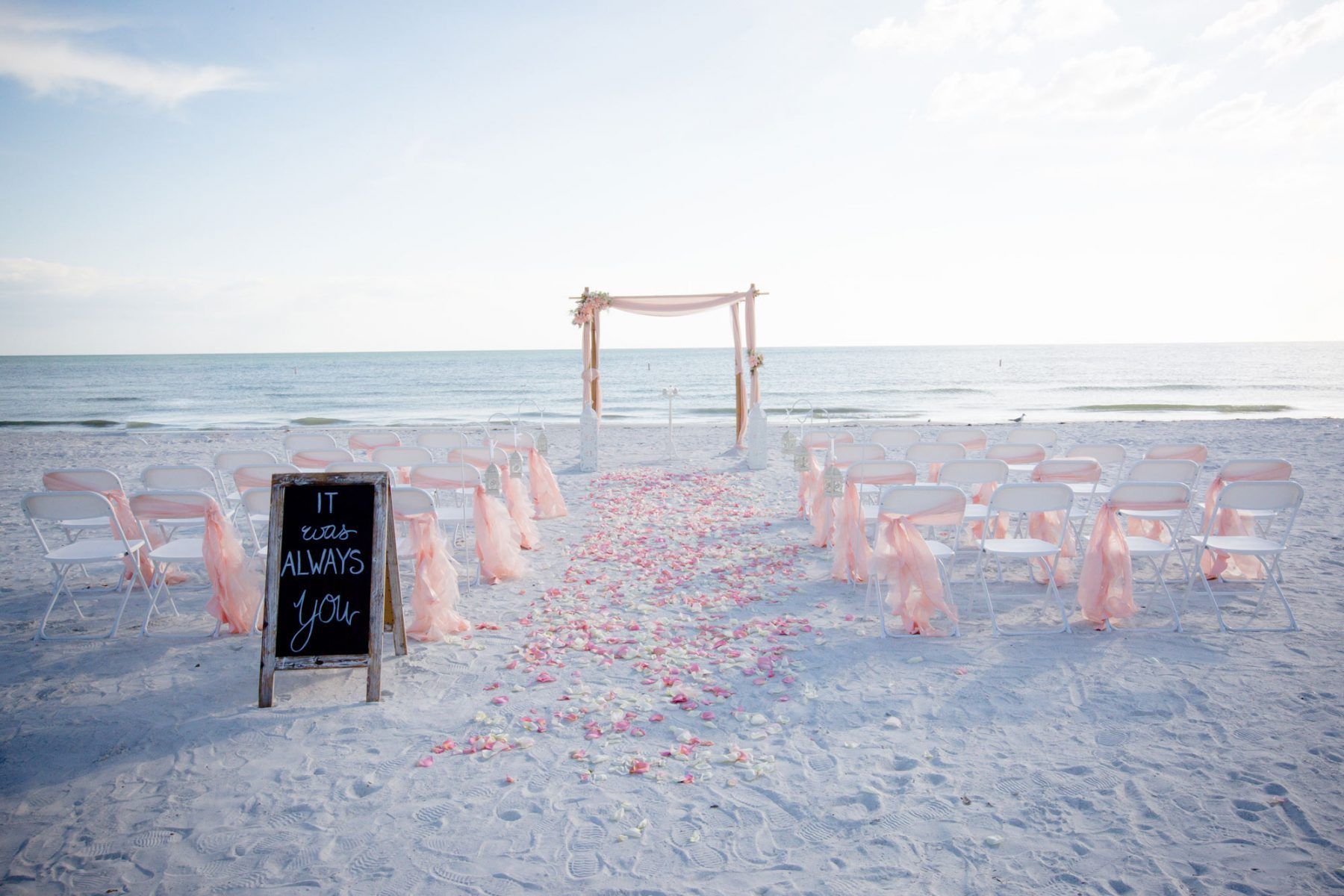 Beach wedding setup: chairs with pink sashes, rose petal aisle, archway, and sign with