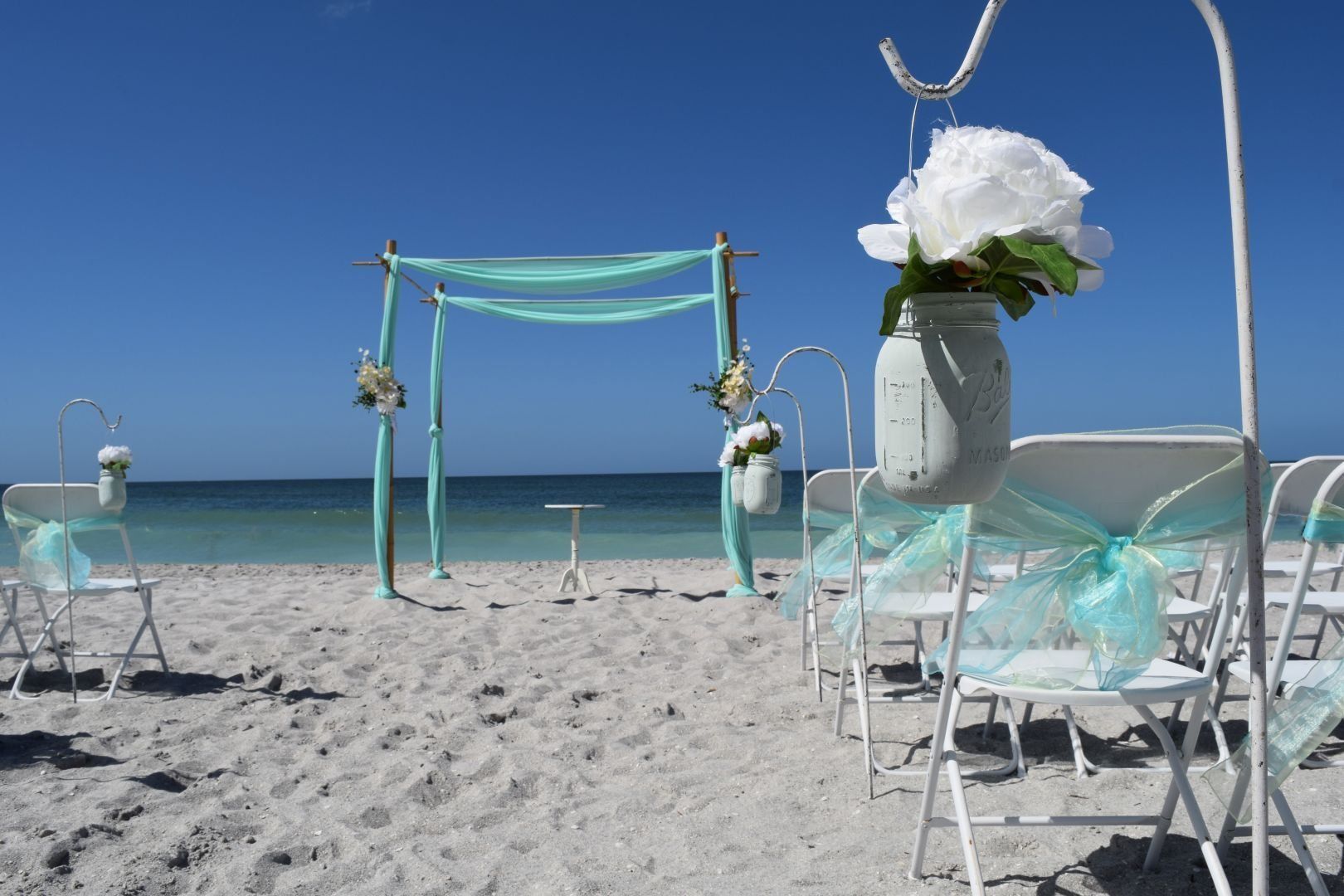 Beach wedding setup with turquoise accents, arch, and flower-filled mason jars.