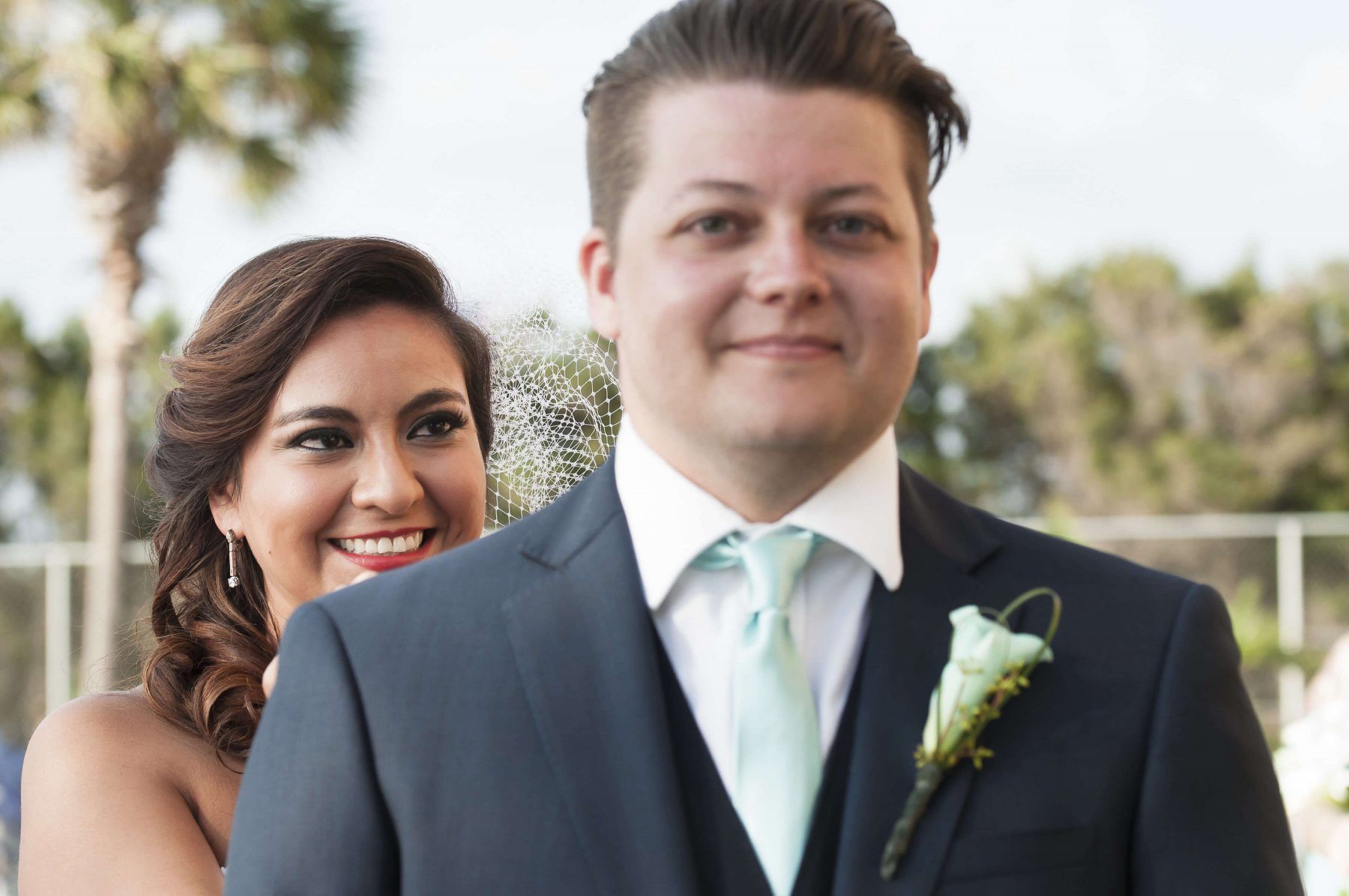 Bride smiles behind groom in a blue suit, outdoor wedding setting.