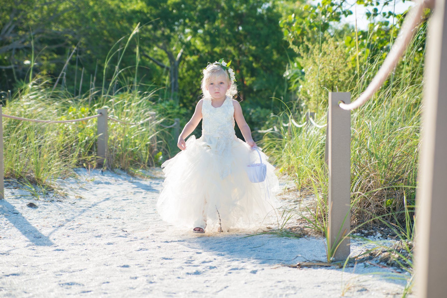 Young girl in white dress carrying a basket walks on a sandy path toward camera.