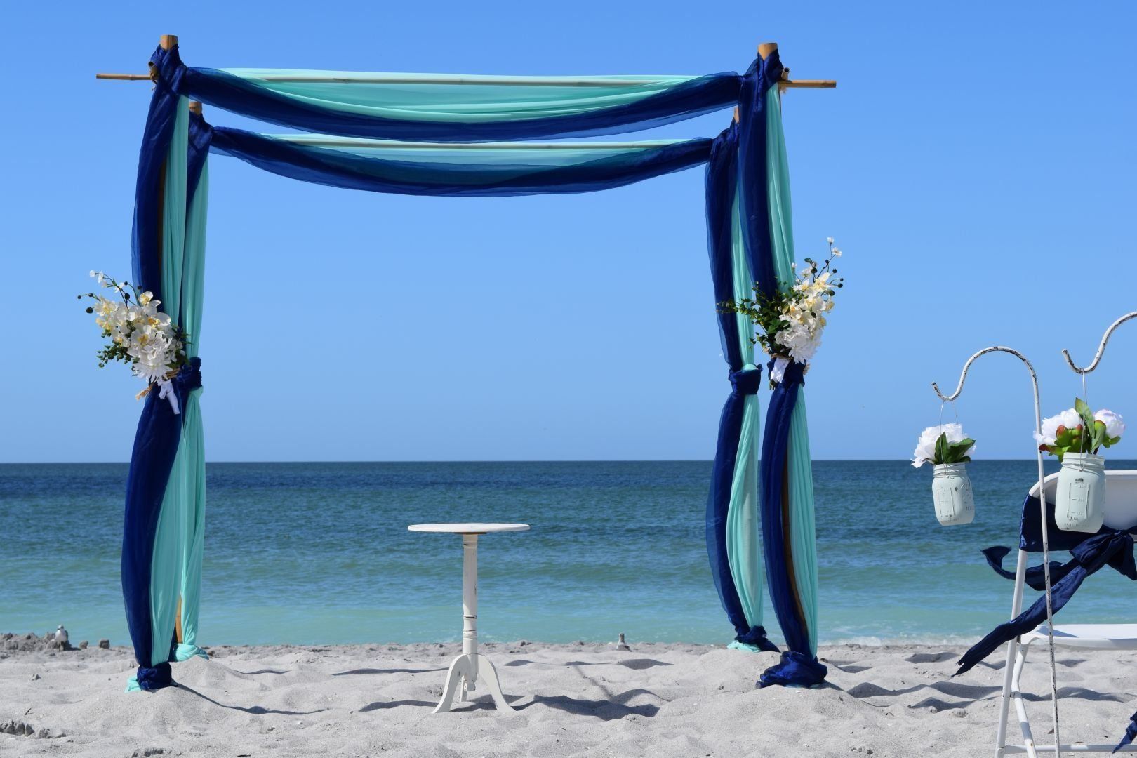 Wedding arch on a beach, blue and teal draping, small table, white flowers, ocean and blue sky in background.