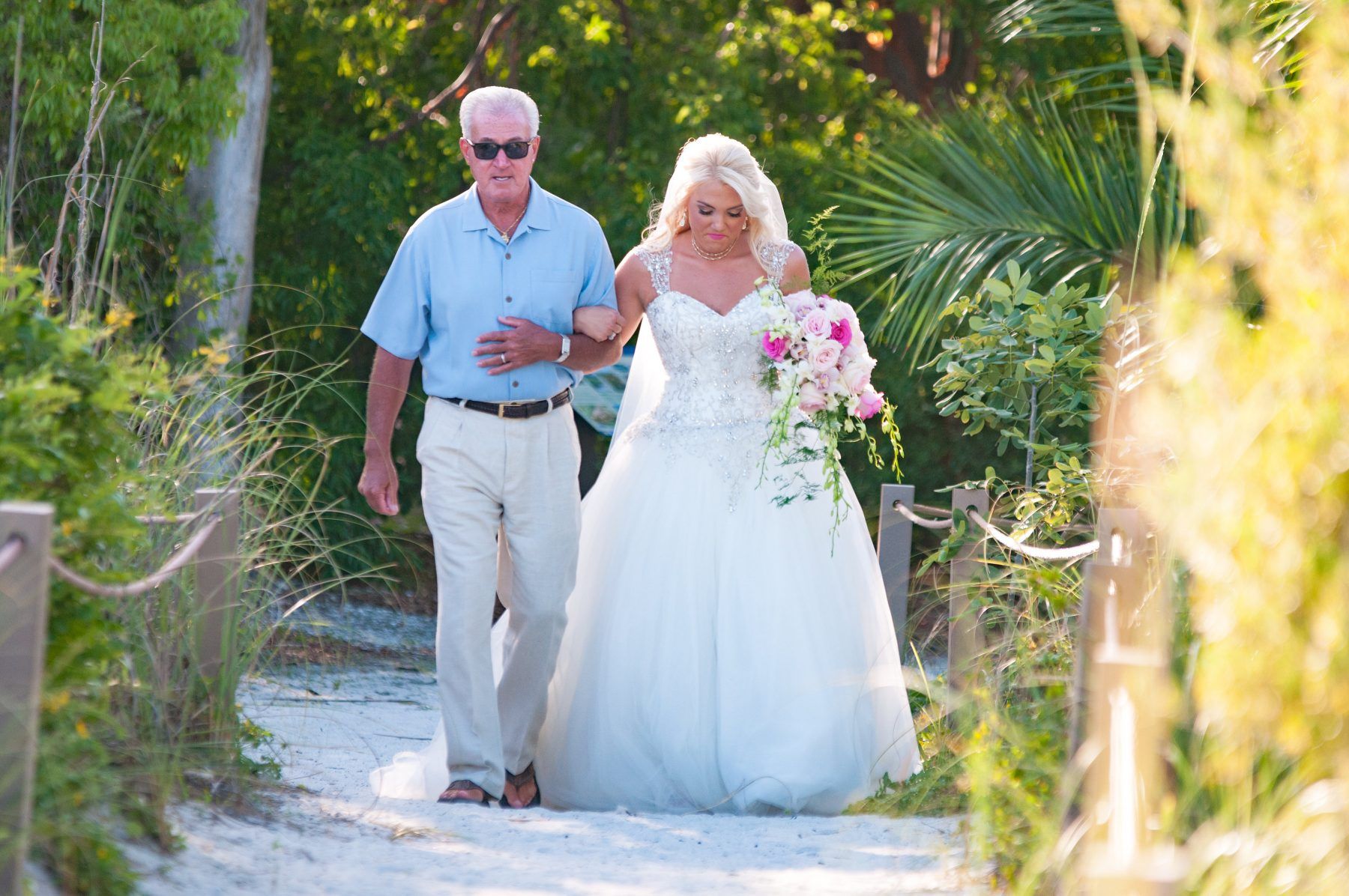 Bride in white dress walks down sandy path with her father, holding a bouquet.