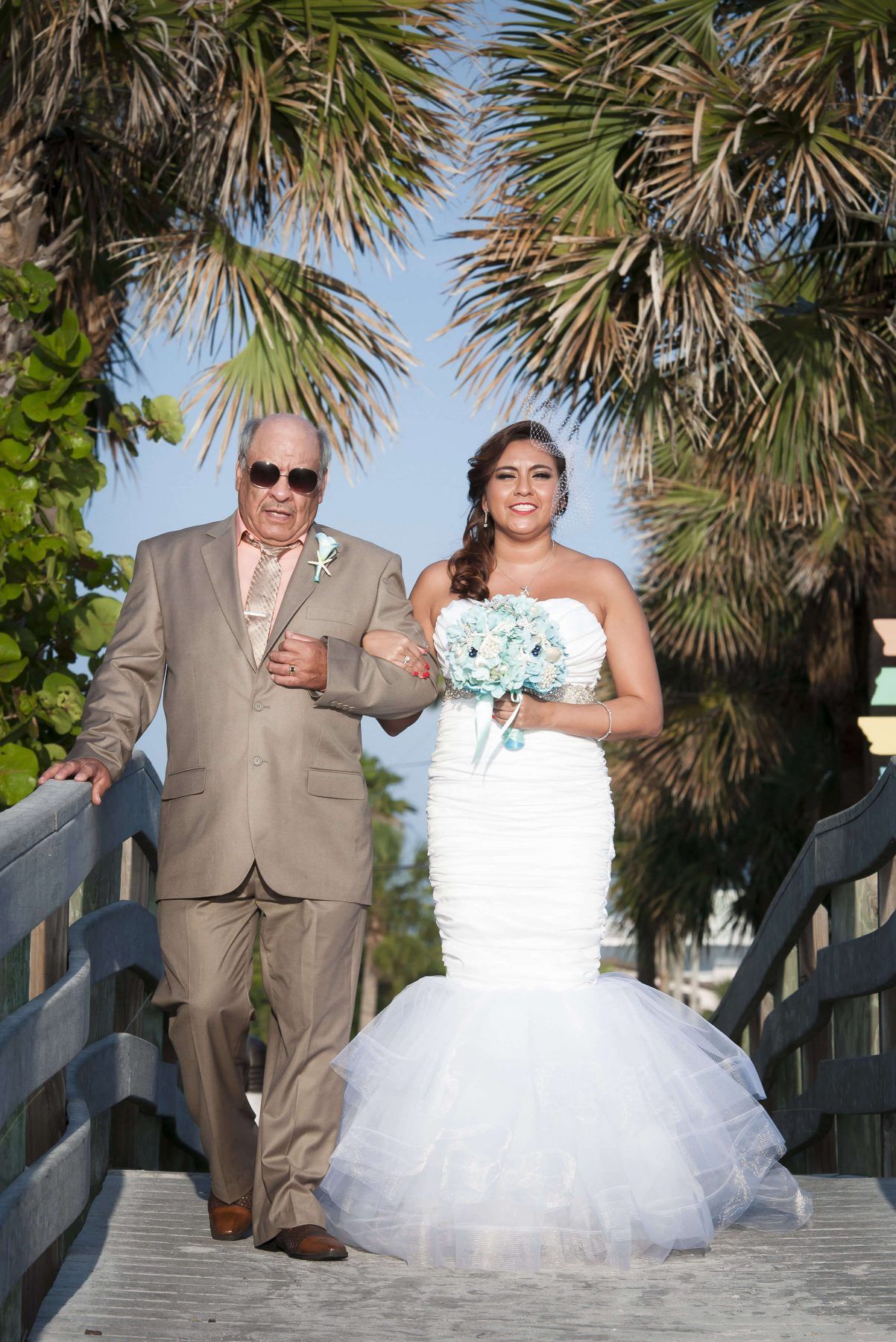 Bride and her father walking on a wooden bridge, palm trees in the background. Bride in white gown, father in suit.