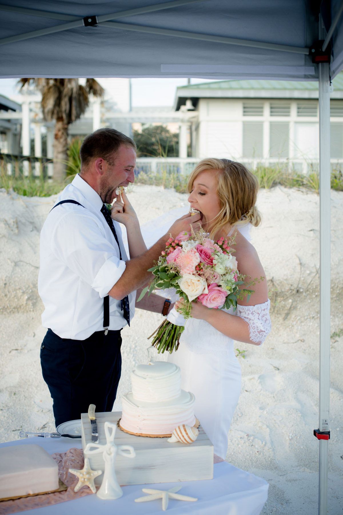 Groom feeds cake to bride on a beach. They laugh. Bride holds bouquet. Wedding cake on table with shells.