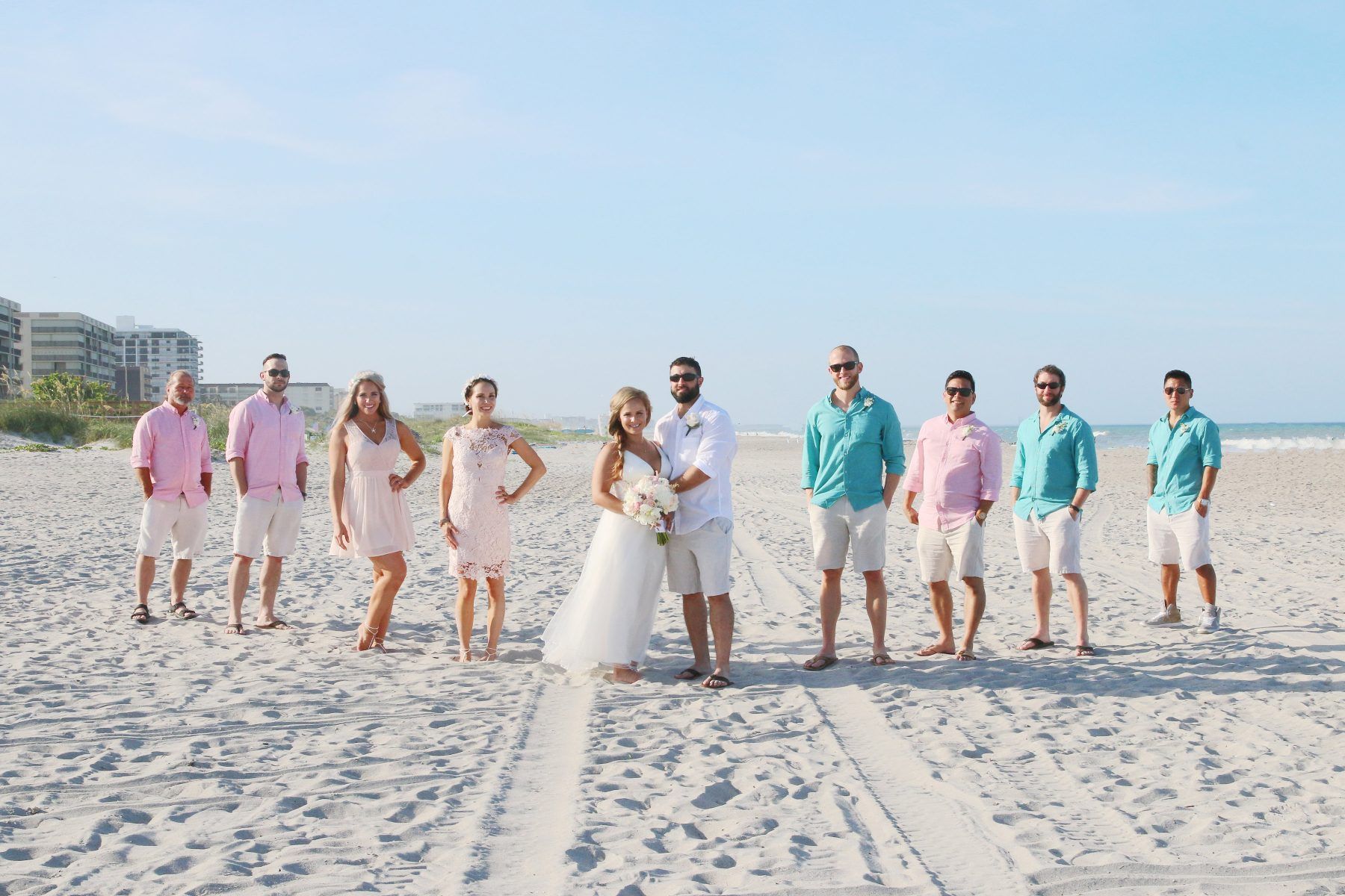 Wedding party on a beach. Bride and groom in center, bridesmaids in pink, groomsmen in teal and pink, all wearing shorts.