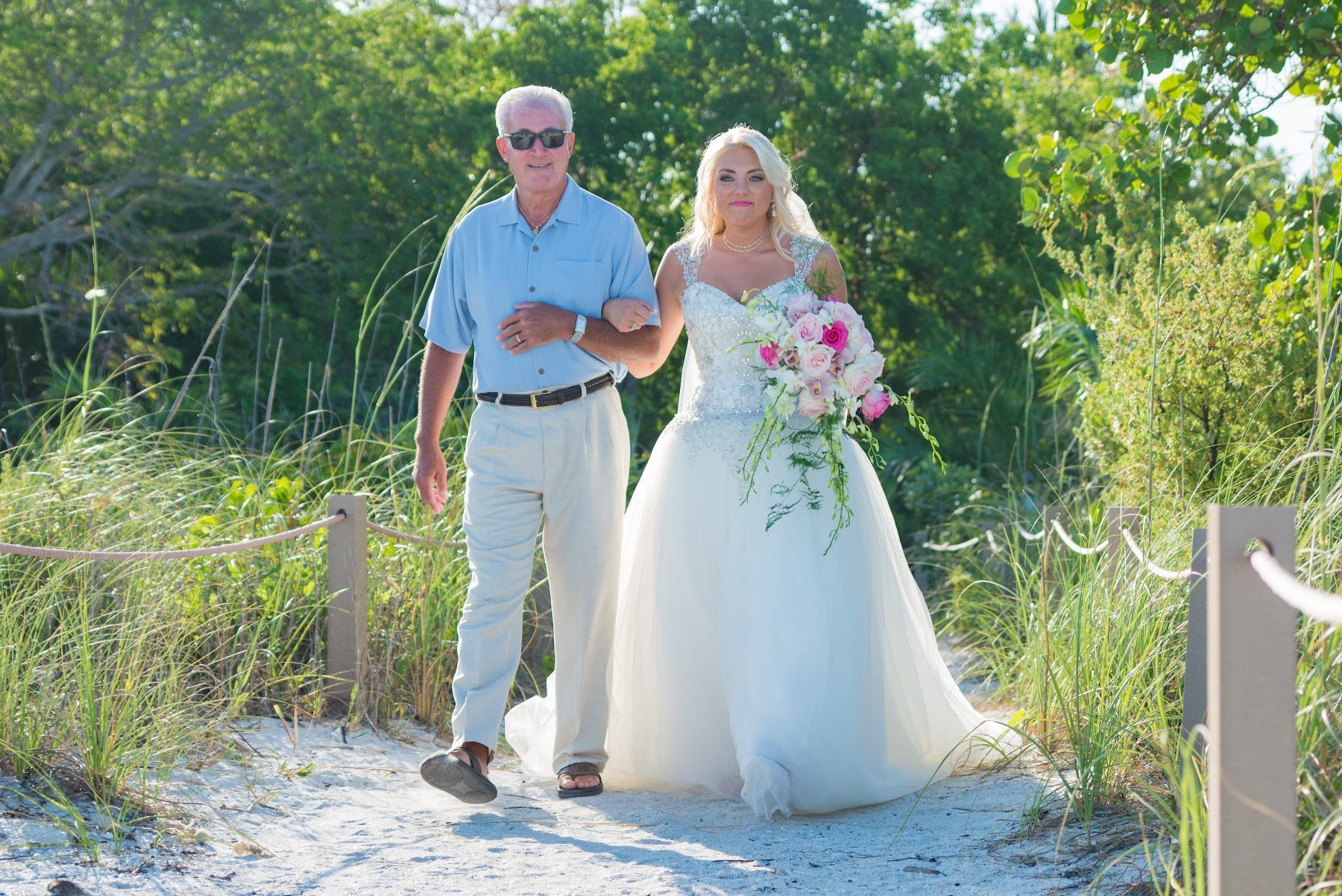 Bride in white gown walks with father along sandy path; beach setting.