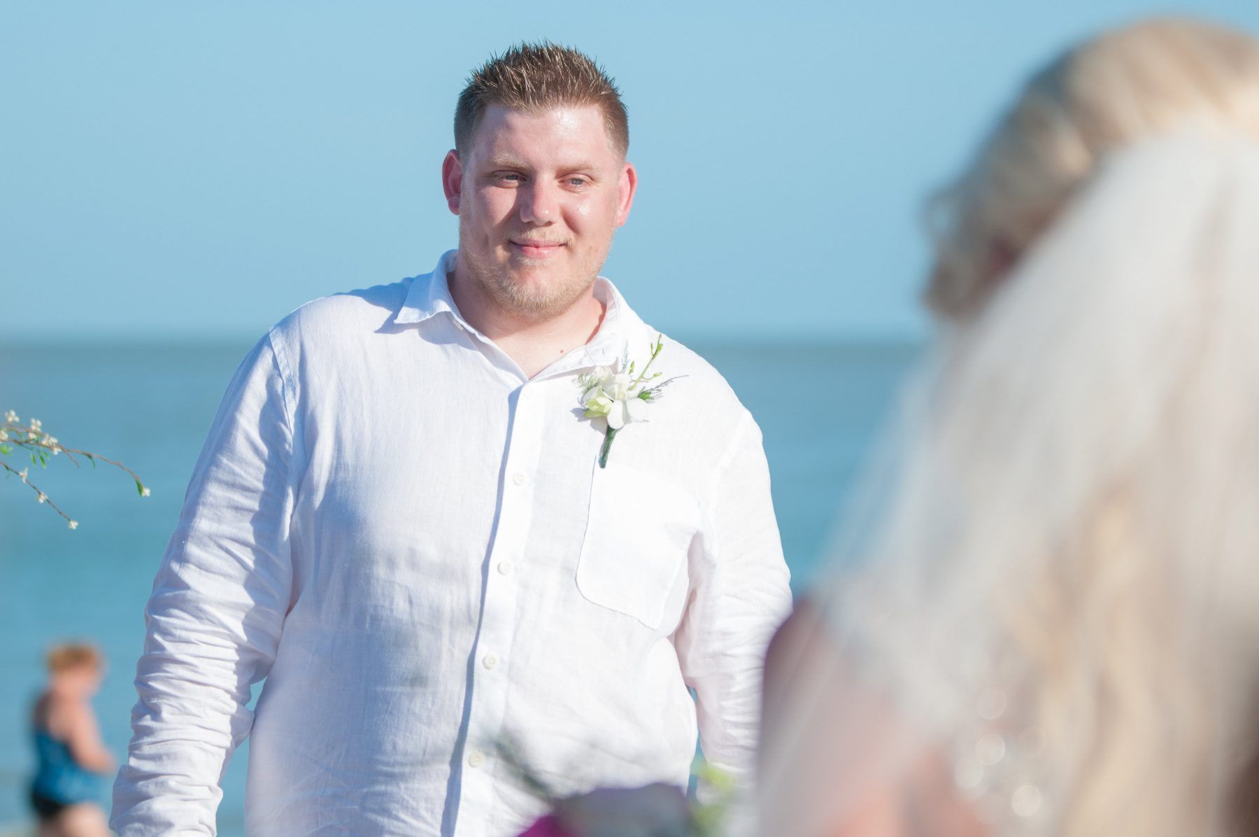 Groom in white shirt smiles, awaiting bride on a beach wedding. Ocean and blue sky background.