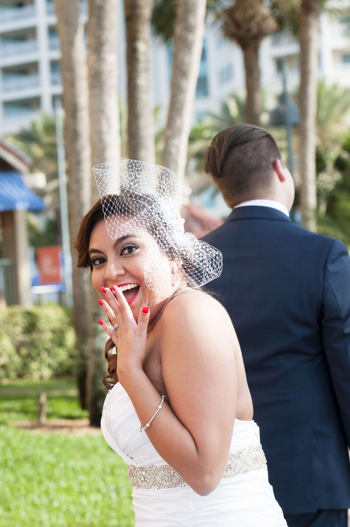 Bride in a white dress, surprised expression, hand over mouth, groom behind her. Outdoor setting, palm trees.