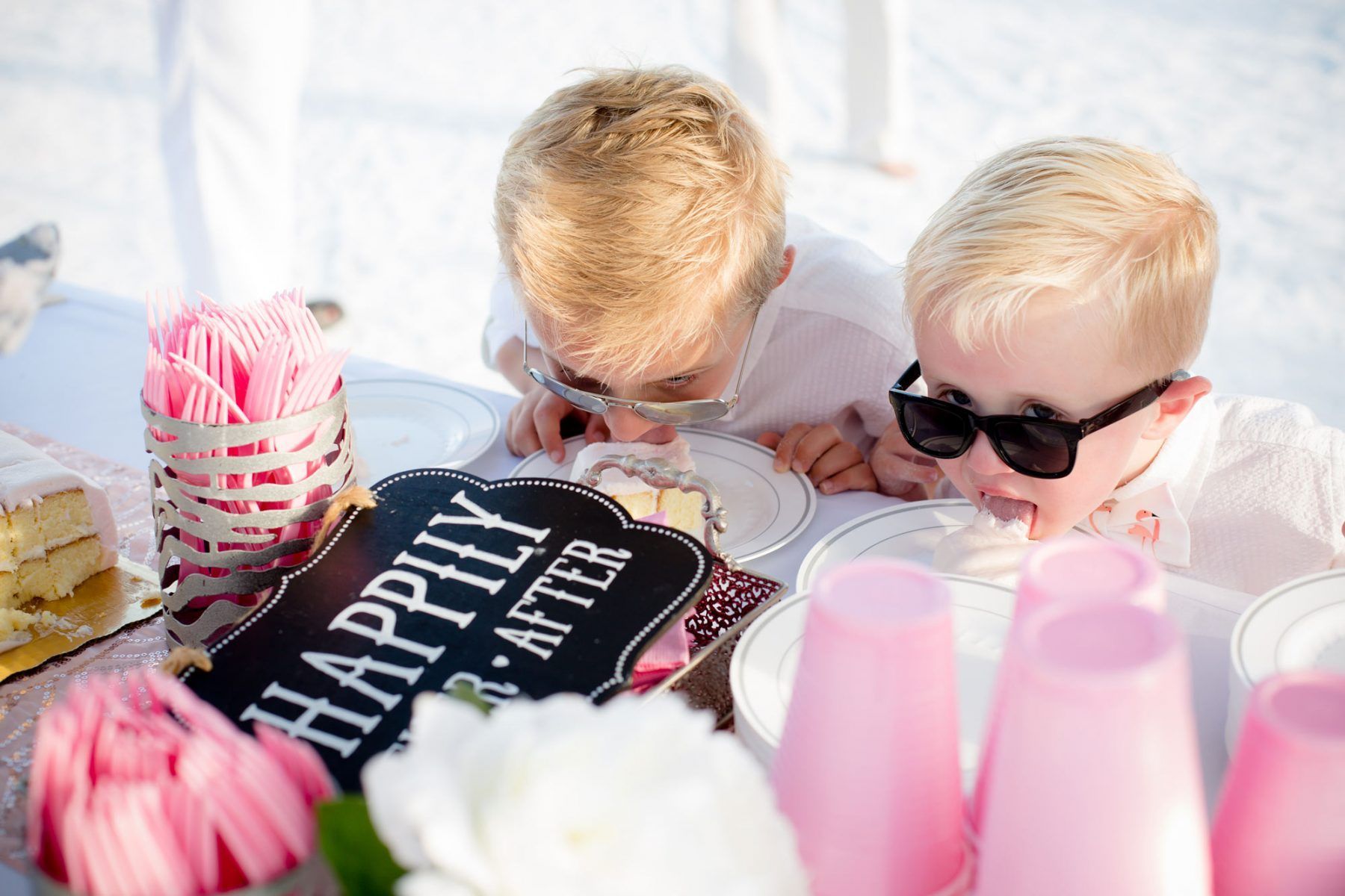 Two children, wearing sunglasses, eating cake off plates at a wedding table with a
