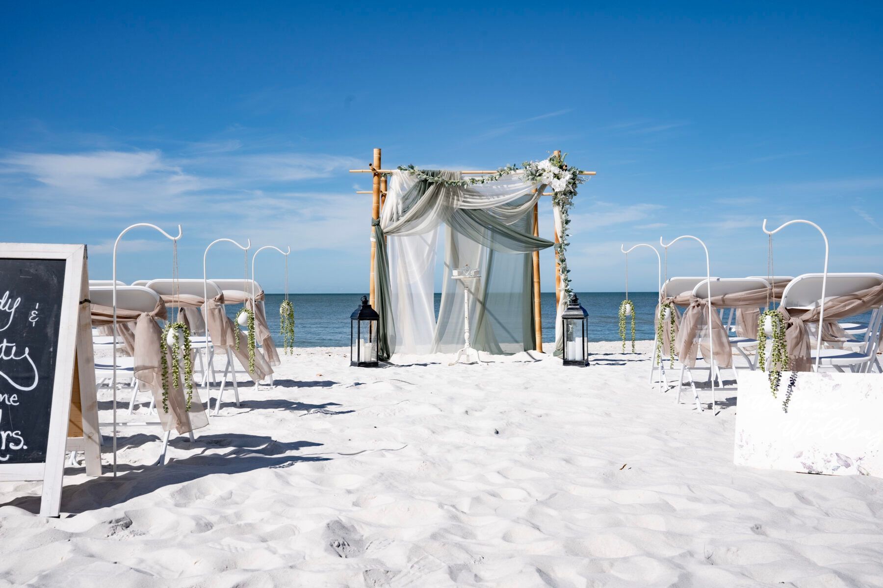 Beach wedding setup with arch, chairs, and lanterns on white sand under blue sky.
