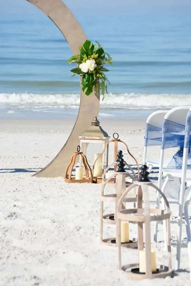 Wedding arch with floral arrangement, lanterns, and chairs on a sandy beach with ocean in the background.