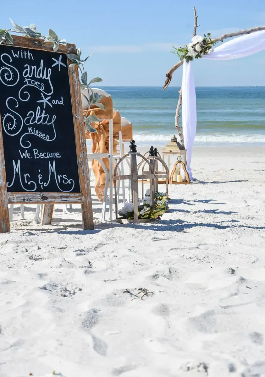 Beach wedding setup with chalkboard sign and floral arch.