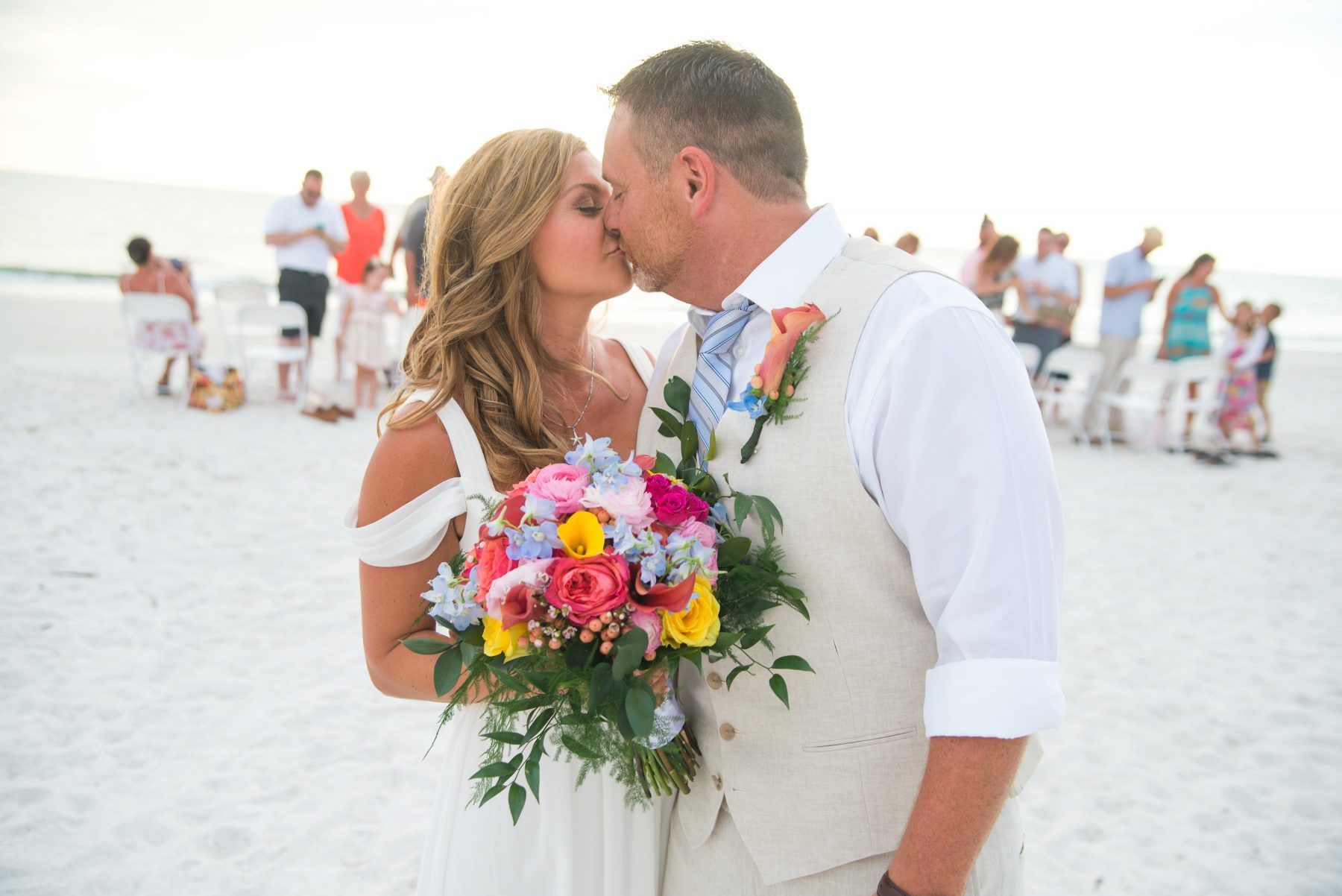 Couple kissing on beach, bride holding colorful bouquet. Guests in background.