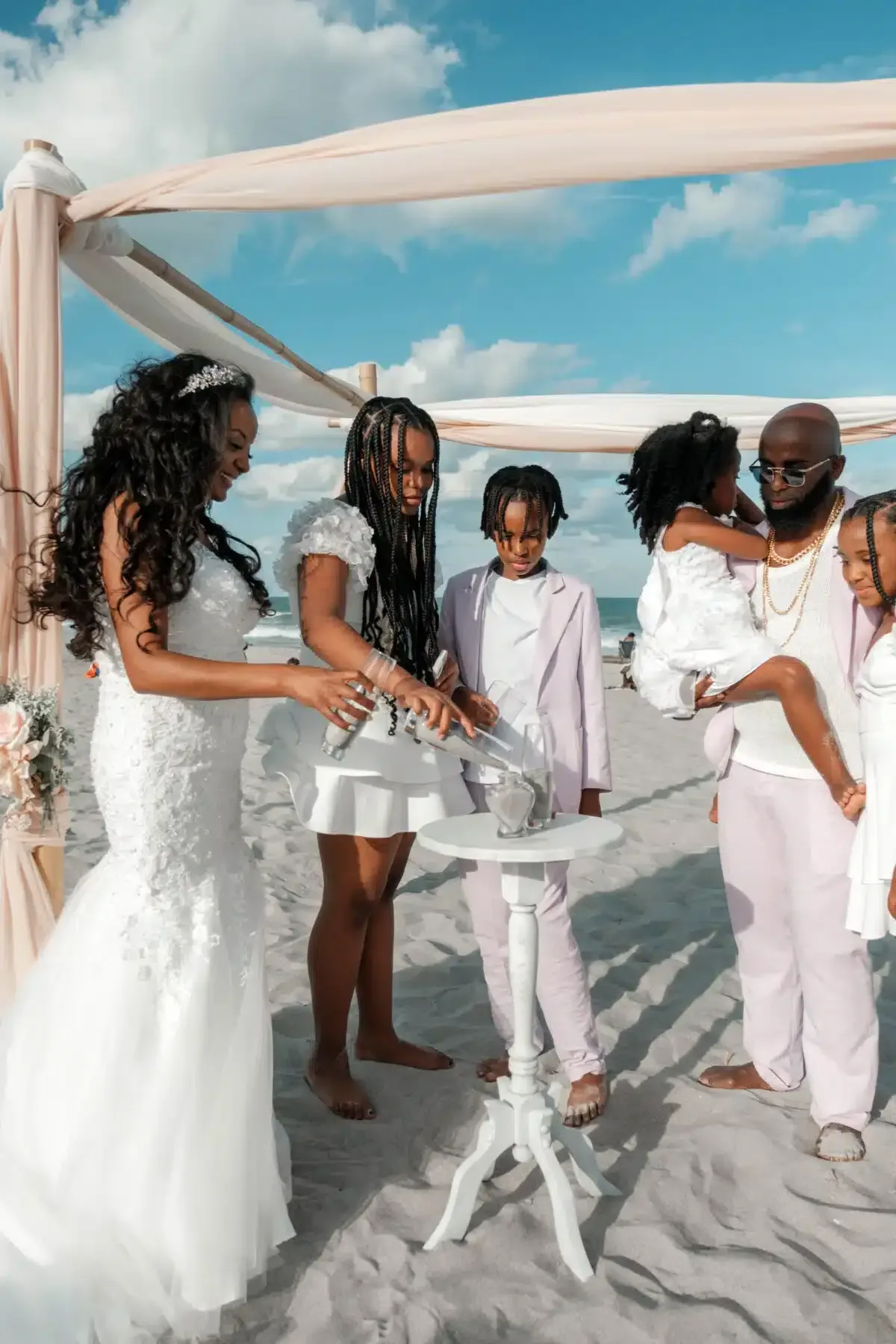 Wedding ceremony on beach; couple and children pouring sand into a vessel under a draped arch.
