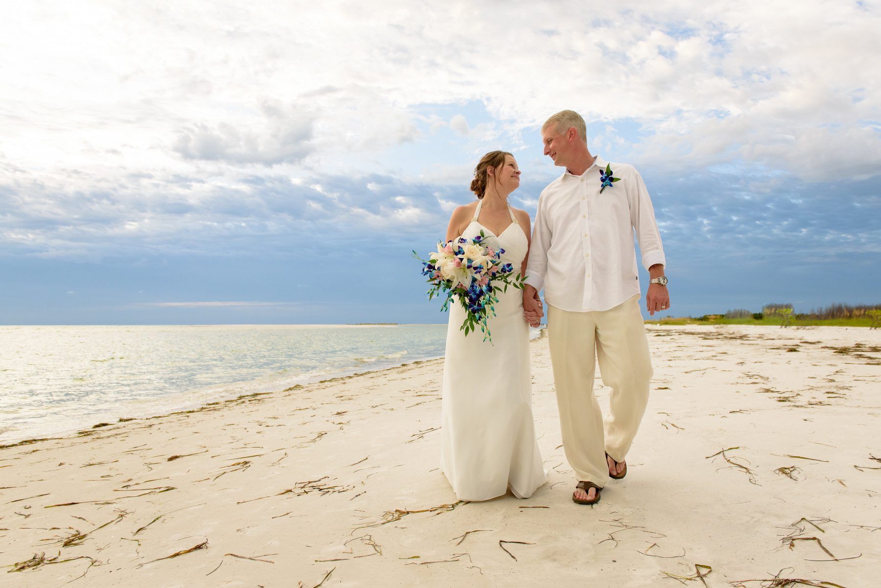 Couple holding hands, walking on beach; bride in white dress, groom in linen pants.