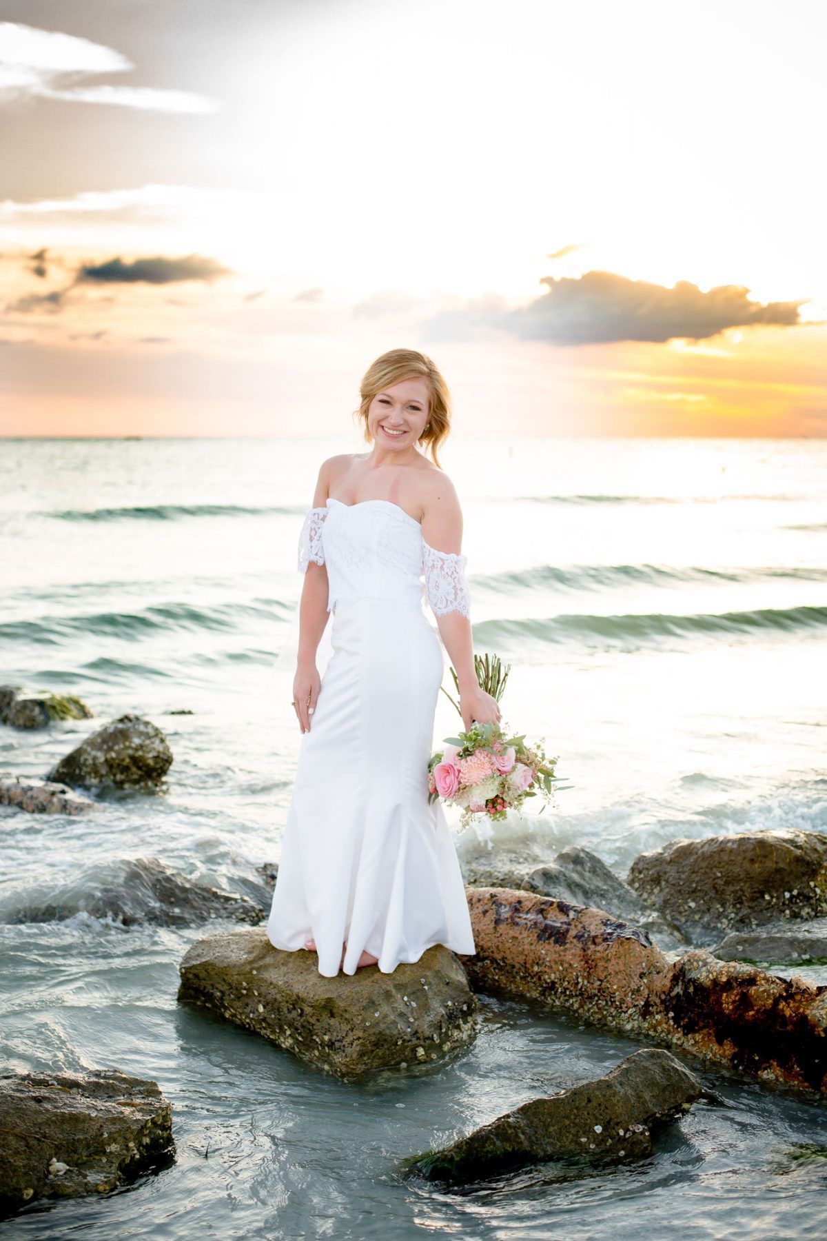 Bride in a white gown stands on a rock at the beach, holding a bouquet, with sunset sky and water.