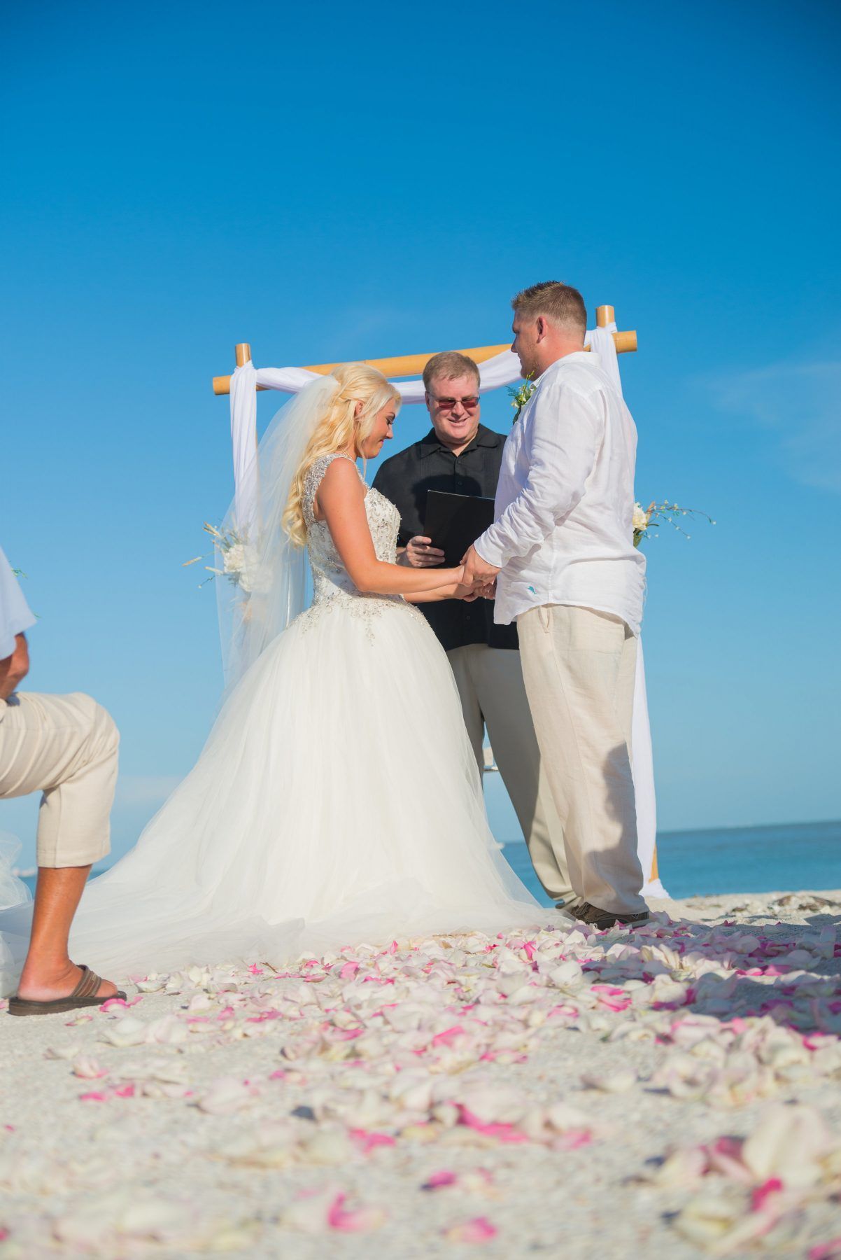 Couple holding hands at beach wedding ceremony under blue sky.