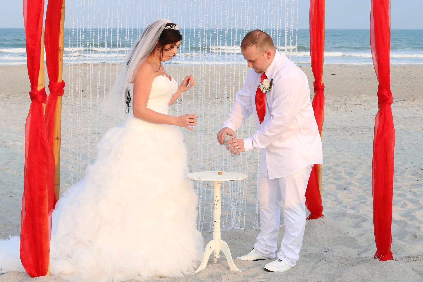 Bride and groom pouring sand during a beach wedding ceremony, red accents, ocean backdrop.