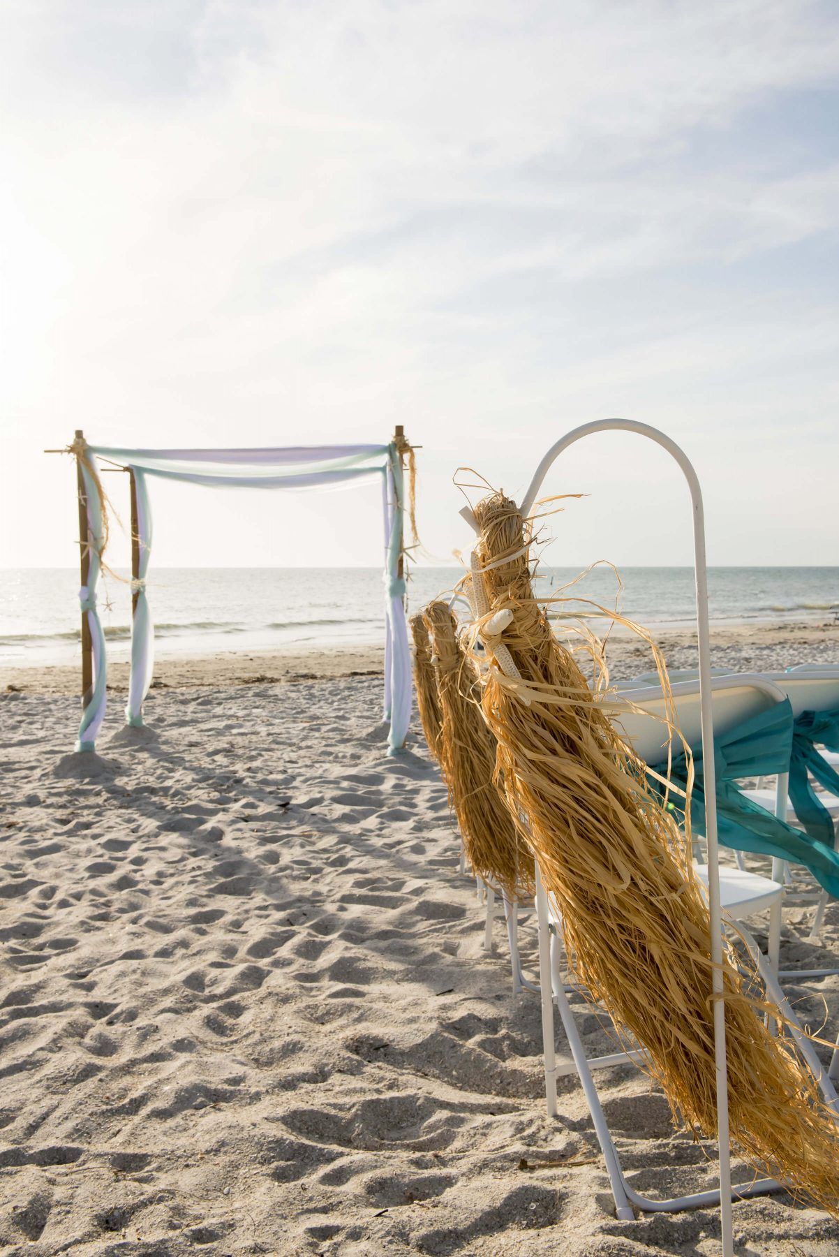 Beach wedding setting with a decorated arch and chairs with bundles of dried grass.